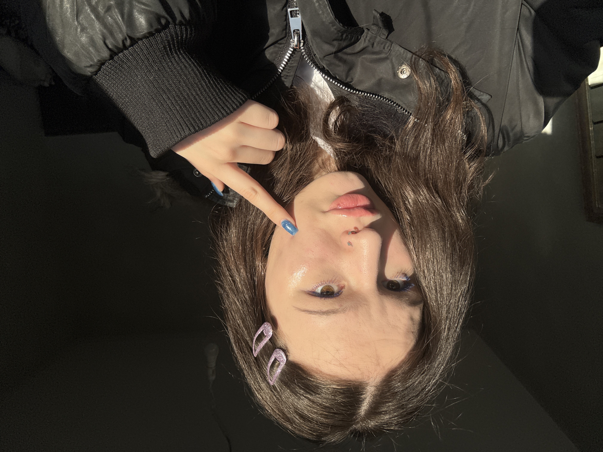 This media file is a high-contrast, sideways-oriented portrait of a young person, likely a woman, positioned from the chest up. She has dark brown hair, styled with two glittery, purple, abstract-shaped hair clips on the right side of her head. She is wearing a black puffer jacket with a visible silver zipper and ribbed black cuffs.
Her left hand is prominent, with the index finger pointing towards her face, specifically near her philtrum piercing. The fingernail on this finger is painted with bright blue glitter nail polish. A subtle silver philtrum piercing is visible on her upper lip. Her makeup includes blue eyeliner accentuating her eyes and light pink lip gloss. Her gaze is directed towards the viewer, with a neutral expression.
The scene is characterized by strong, directional lighting, suggesting direct sunlight streaming into an otherwise dark setting, possibly indoors. This creates significant highlights on her face, hair, and the texture of her jacket, contrasting sharply with the deep shadows that dominate the background. No specific location cues are visible within the image itself to indicate Saint Petersburg, Russia, nor is any text visible. The lighting creates a dramatic and moody atmosphere.