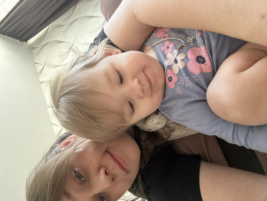 An adult female and a baby/toddler are photographed in an indoor setting, likely a residence in Chaikovsky, Russia. The adult female, with light brown hair, fair skin, and wearing a black top, is partially visible on the left side of the frame, smiling faintly. She appears to be holding the baby/toddler, who is positioned in front of her. The baby/toddler has light blonde hair, fair skin, and is wearing a light blue long-sleeved shirt with a floral pattern of pink and light blue flowers. A necklace with a thin white cord, a small brown cylindrical bead, and a silver-colored cross pendant hangs around the baby's neck. Small red stud earrings are visible in the baby's ears. The baby is looking downwards with a slight pout. The background consists of a light-colored wall with a grey, feather-like patterned wallpaper. Dark brown curtains are visible on the right, and a section of a brown upholstered sofa or chair is visible, suggesting the subjects are seated on it.