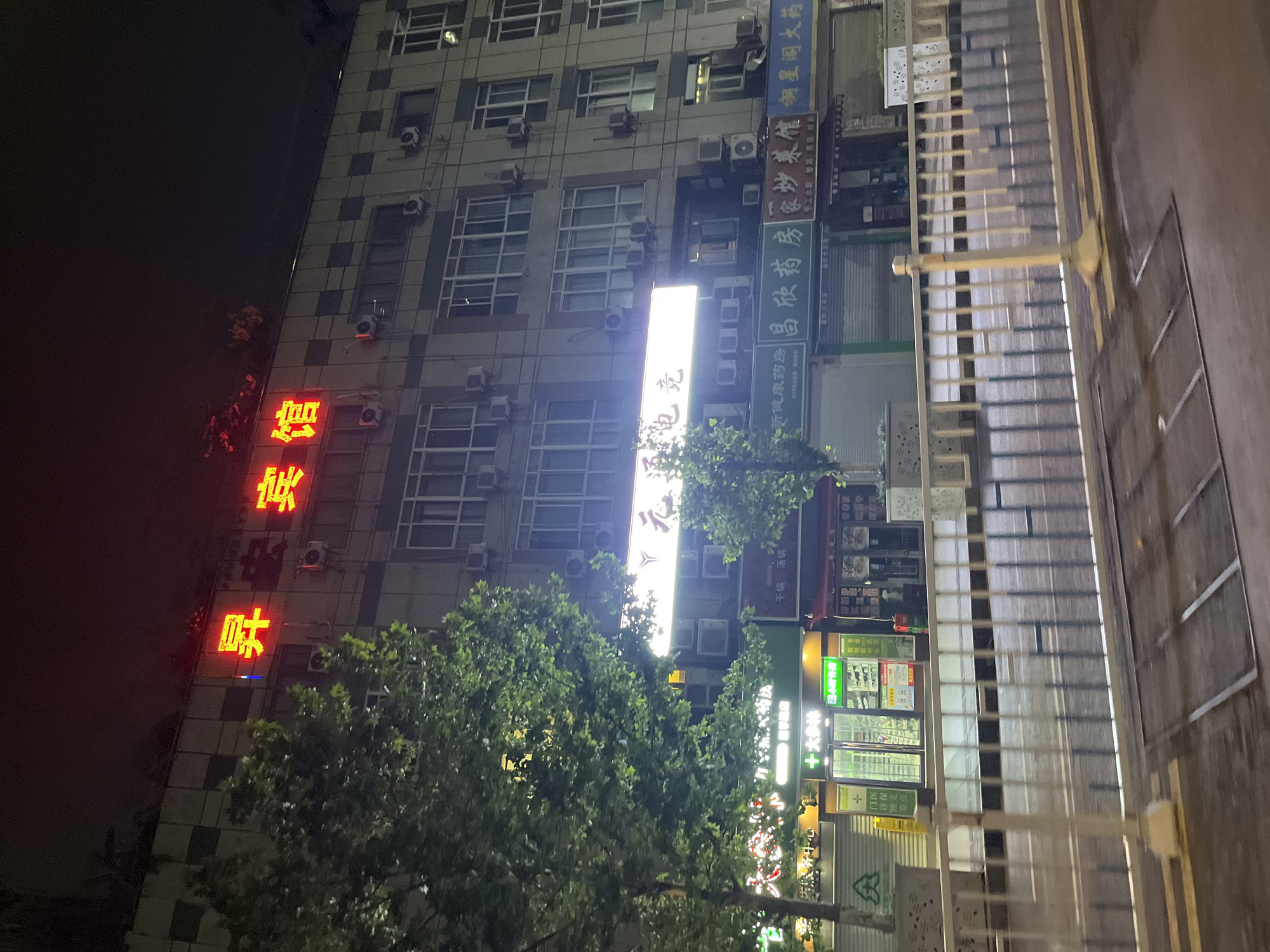 The image shows a multi-story building at night in Jiulongpo District, China. The building has a prominent red neon sign at the top with Chinese characters that read "昇安宾馆" (Sheng An Hotel). Below this sign, a horizontal illuminated sign with Chinese characters "元通电竞" (Yuan Tong E-sports) is visible. Various shop fronts are visible on the ground floor, including a pharmacy with a green illuminated sign featuring a plus symbol and Chinese characters, and other businesses with closed shutters. Air conditioning units are mounted on the exterior walls of the building. A metal railing with decorative elements separates the sidewalk from a road. The street is wet, suggesting recent rain. Trees with green foliage are present on the left and in front of the building. The scene is dimly lit by artificial lights, creating a nighttime ambiance typical of an urban environment. No people are visible in the image.