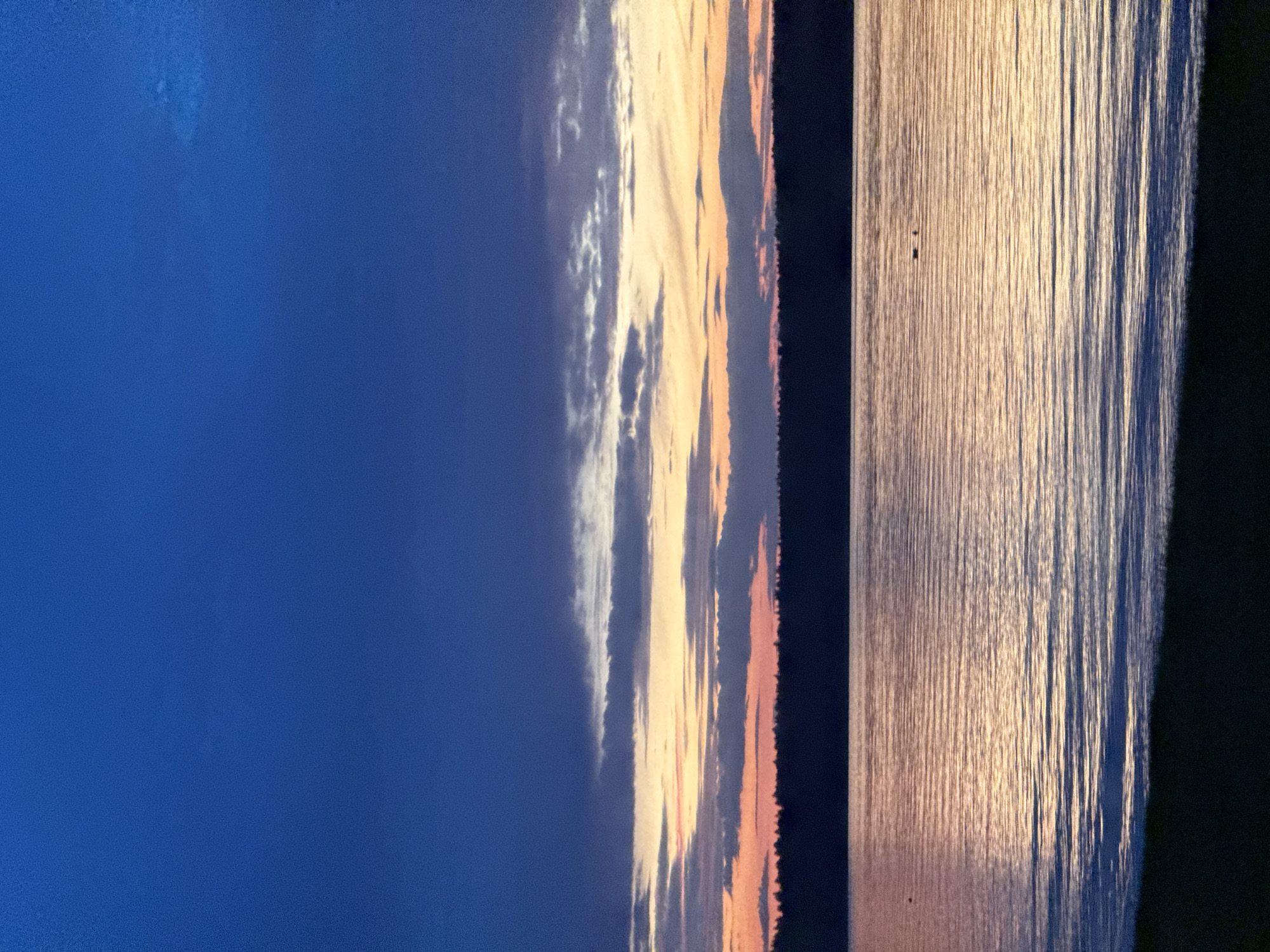 The image depicts a tranquil lake scene at dusk. The sky is a deep, rich blue, transitioning to softer hues of orange and pink where the sun has set below the horizon. Wispy clouds streak across the sky, catching the last rays of sunlight. A dark, silhouetted treeline forms a distinct horizon line above the water. The lake's surface is calm, reflecting the colors of the sky and illuminated by the fading light, creating shimmering patterns of light and shadow. In the foreground, the dark silhouette of a grassy shore is visible. The overall atmosphere is peaceful and serene. The image is not geolocated to Votkinsk, Russia; it shows a generic lake scene.