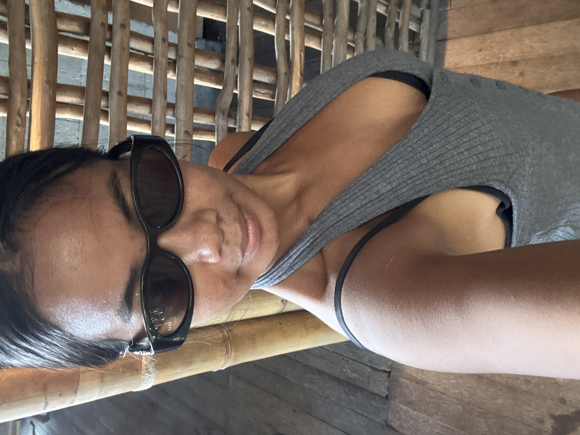 A woman with dark hair, wearing sunglasses, a grey tank top, and a black bra, takes a selfie. She is standing in front of a bamboo screen and a bamboo pole, with wooden flooring beneath her. The setting appears to be a resort or outdoor dining area in North Kuta, Indonesia.