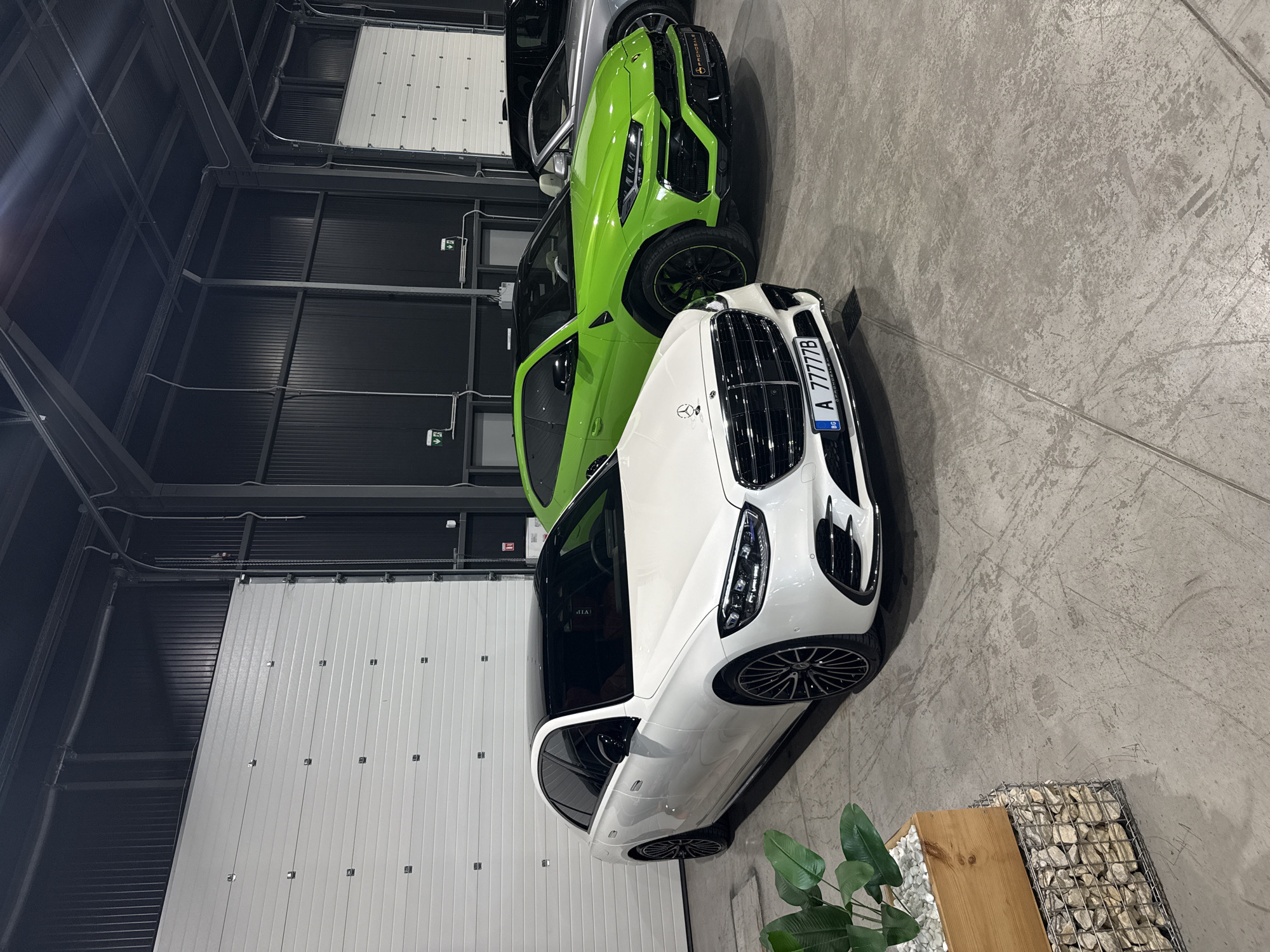 The image shows a garage or showroom in Plovdiv, Bulgaria. There are three luxury cars parked inside. The car closest to the camera is a white Mercedes-Benz with a Bulgarian license plate "A 77777B." Next to it is a green Lamborghini SUV. Another, likely luxury, car is partially visible behind the Lamborghini. A garage door is visible on the left. A small wooden table with a plant is located in the lower right corner. The floor is concrete. The building has metal walls and ceiling.