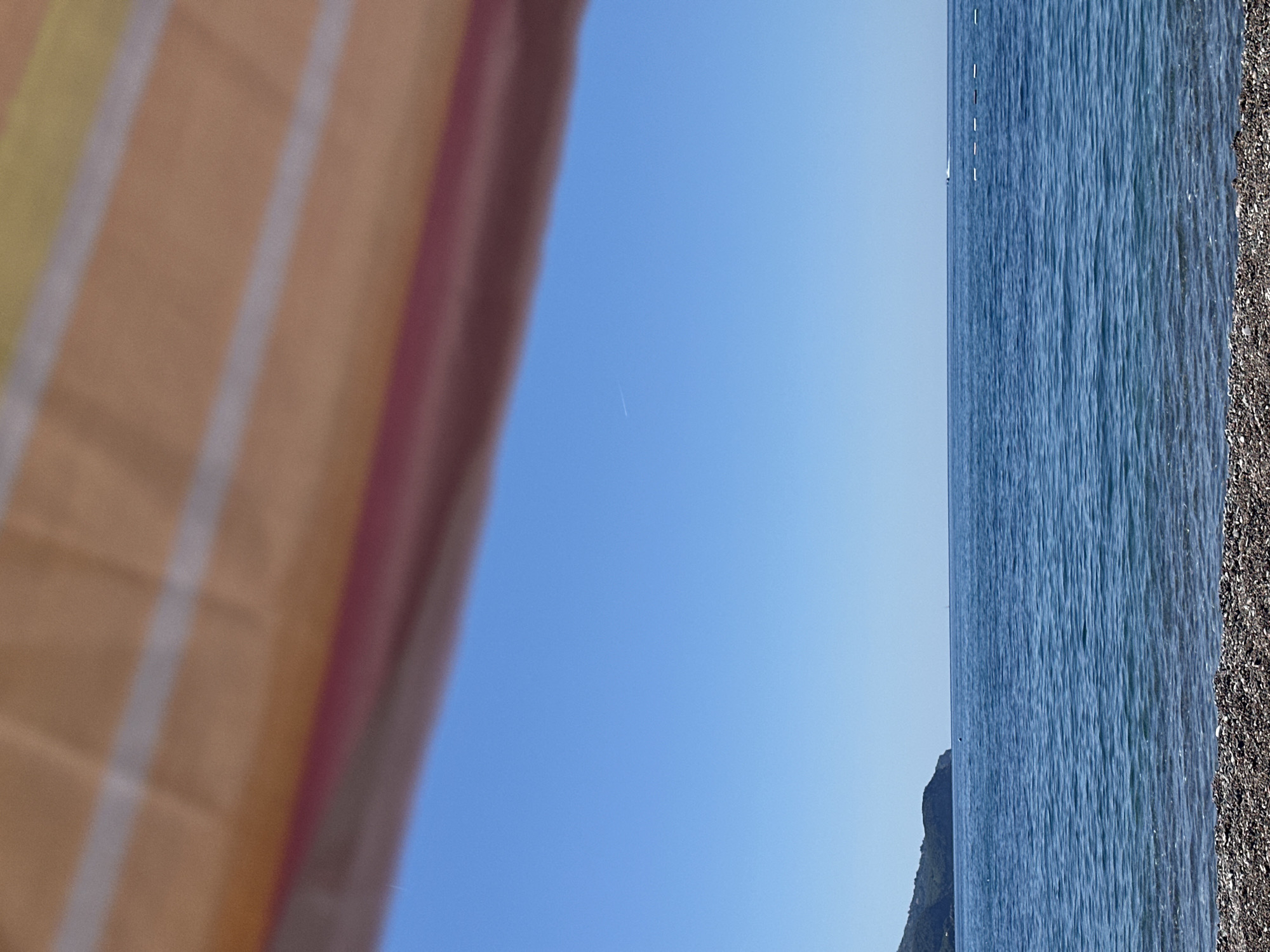 The image presents a view from a beach, potentially in Boreti, Montenegro, encompassing a section of the beach, the sea, the sky, and a partial view of what seems to be a beach umbrella or towel in the foreground. The sea appears calm with subtle wave patterns, transitioning to a pebble beach. The sky is clear blue with a faint contrail suggesting an aircraft's passage. A partially obscured object, possibly a small rock formation, is visible near the waterline.