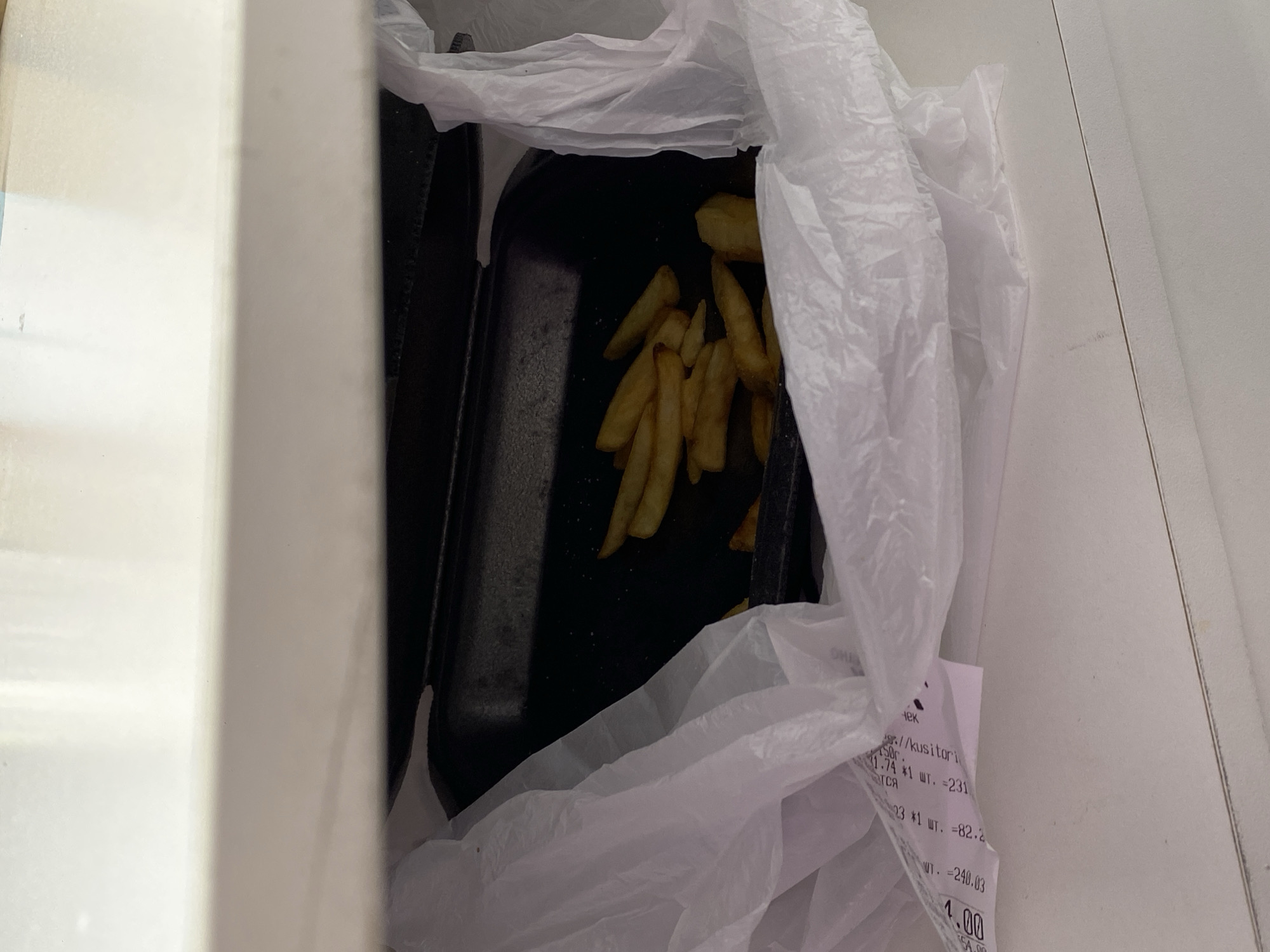This media file depicts a close-up, overhead view of a white plastic bag containing food, specifically french fries, inside a black disposable container. The bag and container are placed within a white box or cooler. A crumpled receipt is partially visible, sticking out from the bag. The receipt shows Russian text and numbers, including "1.74 *1 шт. =231", "23 *1 шт. =82.2", "WT.=240.03", and "4.00". The overall scene suggests a takeaway food order. The lighting appears to be indoor, possibly natural light from above, and there are no indications of the time of day or weather. The location context is Uchaly, Russia.