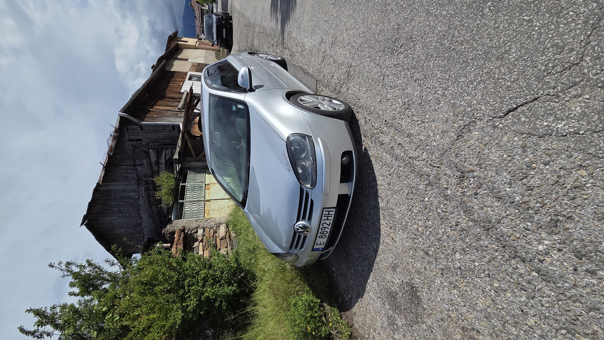 A silver Volkswagen Golf is parked on the side of an asphalt road in Bania, Bulgaria, under a bright, partly cloudy sky. The car, likely a Mk5 model, is angled towards the right side of the frame, with the front visible. Strong sunlight creates a distinct shadow of the car on the road surface.
The front license plate of the Volkswagen displays "E 8892 HH". A small, unreadable graphic sticker is present on the front hood/headlight, and a purple sticker with the number "019" is visible on the passenger side mirror.
To the left of the parked car stands an old, two-story wooden building with a weathered facade and a corrugated metal roof, suggesting traditional rural architecture. Green bushes and grass grow along the edge of the road, partially obscuring a stone wall in front of the building. In the background, further down the road, another dark-colored car is parked. The asphalt road shows signs of wear and minor cracking. The scene depicts a quiet daytime setting in a village or small town. No individuals are visible.