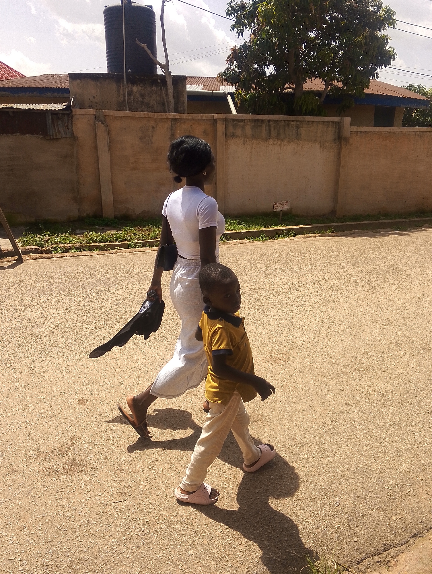 In Jos, Nigeria, on a sunny day, a woman and a young boy walk side-by-side on a paved road. The woman, with her back to the camera, wears a white short-sleeved shirt and light-colored pants. She carries a black bag over her shoulder and holds a black item in her right hand. The boy, to her right and slightly in front, is dressed in a yellow polo shirt and light-colored pants. He looks to his left. A wall runs along the background, with a large water tank visible on top of it. Trees and buildings are also present in the distance. The road is textured, with shadows cast by the individuals and a nearby object on the left.