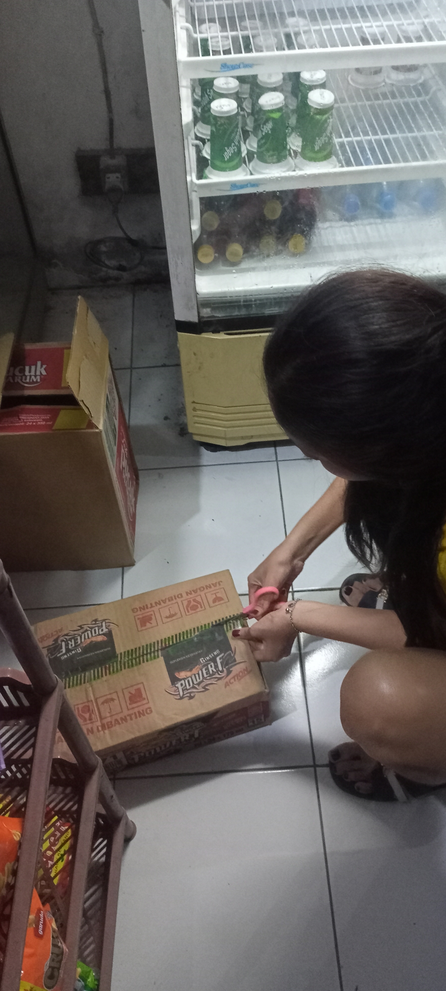 In Gempolklutuk, Indonesia, a person is using pink scissors to open a cardboard box labeled "Power F Ginseng Action." The box is on the floor, next to another cardboard box with "ARUM" visible on it. A white and yellow refrigerator filled with green cans and yellow-capped bottles is to the right. The floor is tiled in gray.
