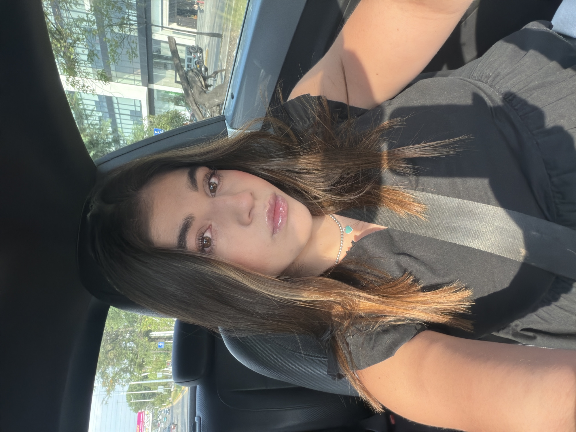 A young woman is depicted in a selfie taken from inside a car, likely in the passenger seat, as indicated by the visible seatbelt across her chest. She has long, dark brown hair with lighter highlights, styled to fall over her shoulders. Her face is turned slightly towards the camera, revealing brown eyes, defined eyebrows, and glossy lips. She is wearing a black top and a silver beaded necklace with a small turquoise heart-shaped pendant.
The setting is inside a car, with elements like the car seat, dashboard, and window frame visible. Through the car window, an outdoor scene is visible. Bright sunlight illuminates the woman and parts of the car interior, suggesting it is daytime with clear weather. Outside, modern buildings with large glass facades reflect the surrounding environment, including trees. A road or street is visible in the distance, with other vehicles and lush green trees lining the street. A blue street sign is partially visible, but its text is unreadable. The overall scene suggests an urban or suburban environment. The activity is a self-portrait taken while traveling or parked in a car. The location context provided for the image is San Bernardino Tlaxcalancingo, Mexico.