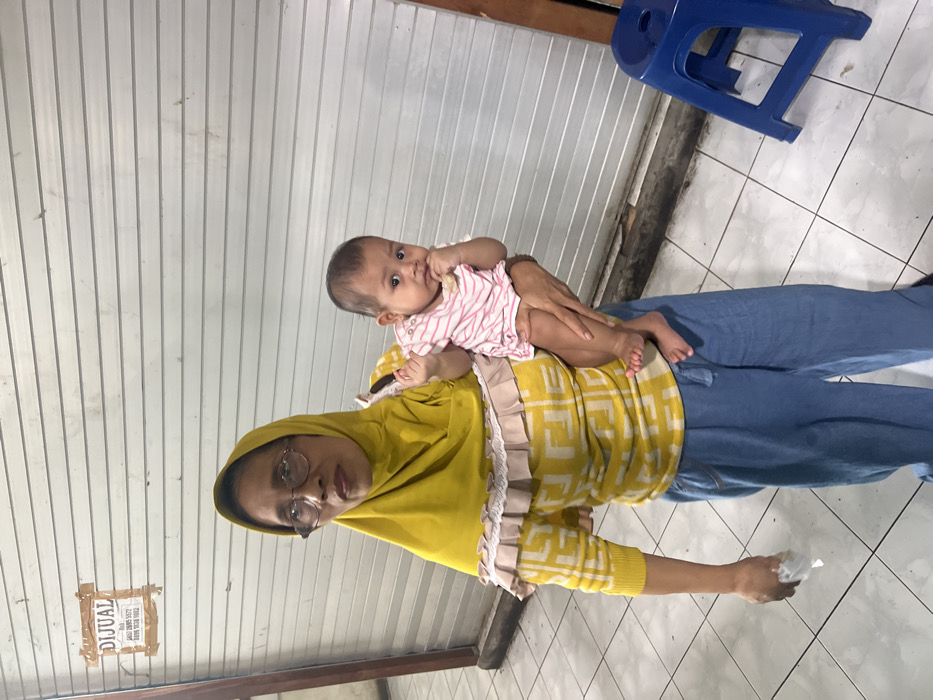 In Nganguk, Indonesia, an adult woman holds an infant against a backdrop of a closed, corrugated metal roll-down shutter. The woman, appearing middle-aged or elderly, wears a yellow hijab, glasses, a yellow long-sleeved shirt with a repeating "F" pattern, and light blue pants. She holds a crumpled white object in her left hand. The infant, clad in a pink and white striped shirt, is held on the woman's left hip, facing the viewer, with its right hand near its mouth, holding a small light-colored object. A "DIJUAL" (FOR SALE) sign, handwritten in red, is taped to the shutter above the woman's head, displaying text and phone numbers. The floor is composed of white tiles, and a blue plastic stool is partially visible in the lower right corner of the frame.