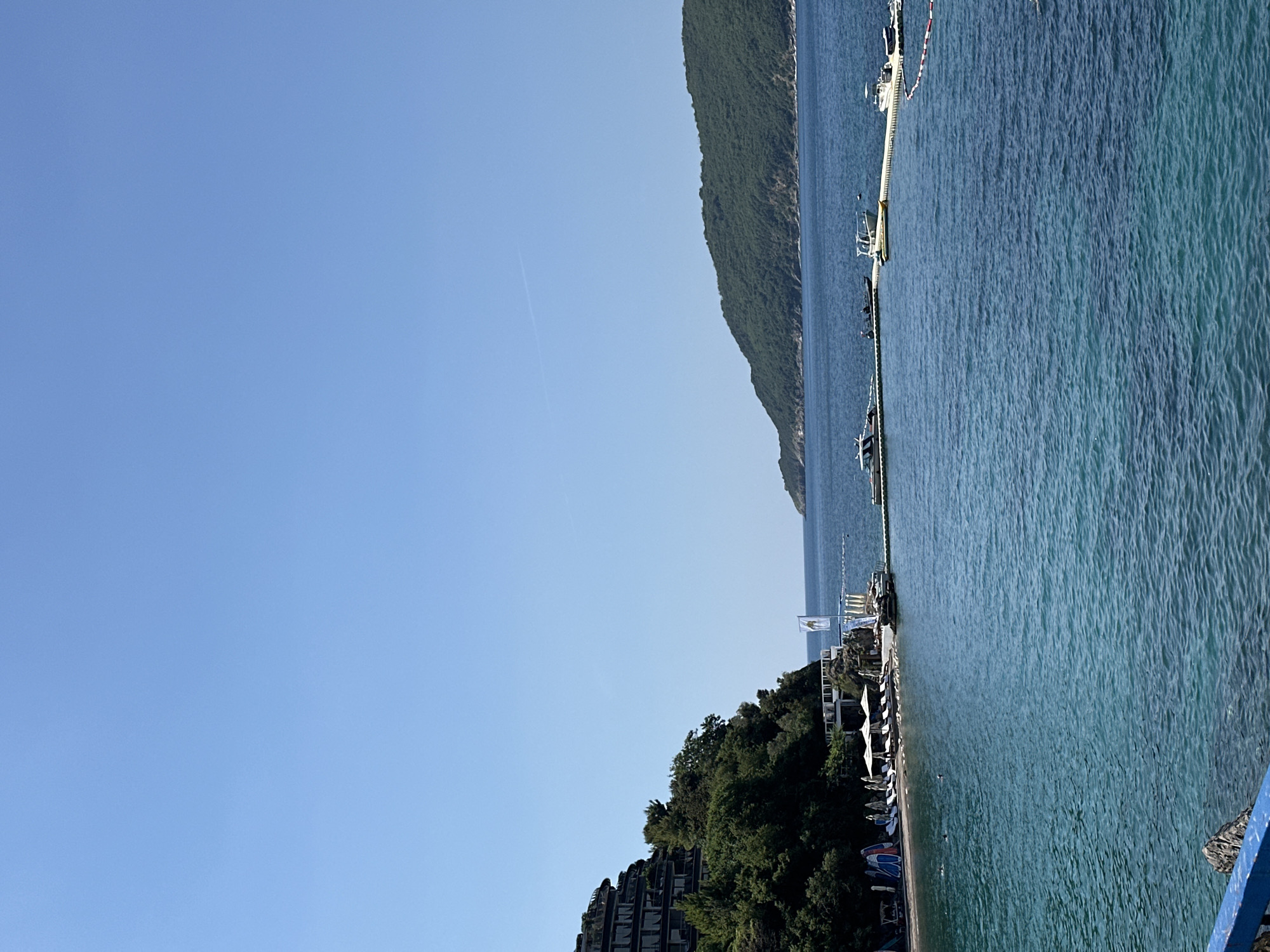 The image shows a view of the sea and a shoreline with a building and vegetation in Boreti, Montenegro. In the foreground, the clear, turquoise water has ripples and shows a gradient of blue. In the mid-ground, a white, floating pontoon extends into the sea, supporting several boats. On the shore, there is a beach with lounge chairs and umbrellas, and a white building with a flag. Behind the beach, a lush green hillside rises, topped by a multi-story building with balconies. In the background, a green, tree-covered island or peninsula juts out into the blue sea, with a faint white contrail visible in the clear blue sky above.