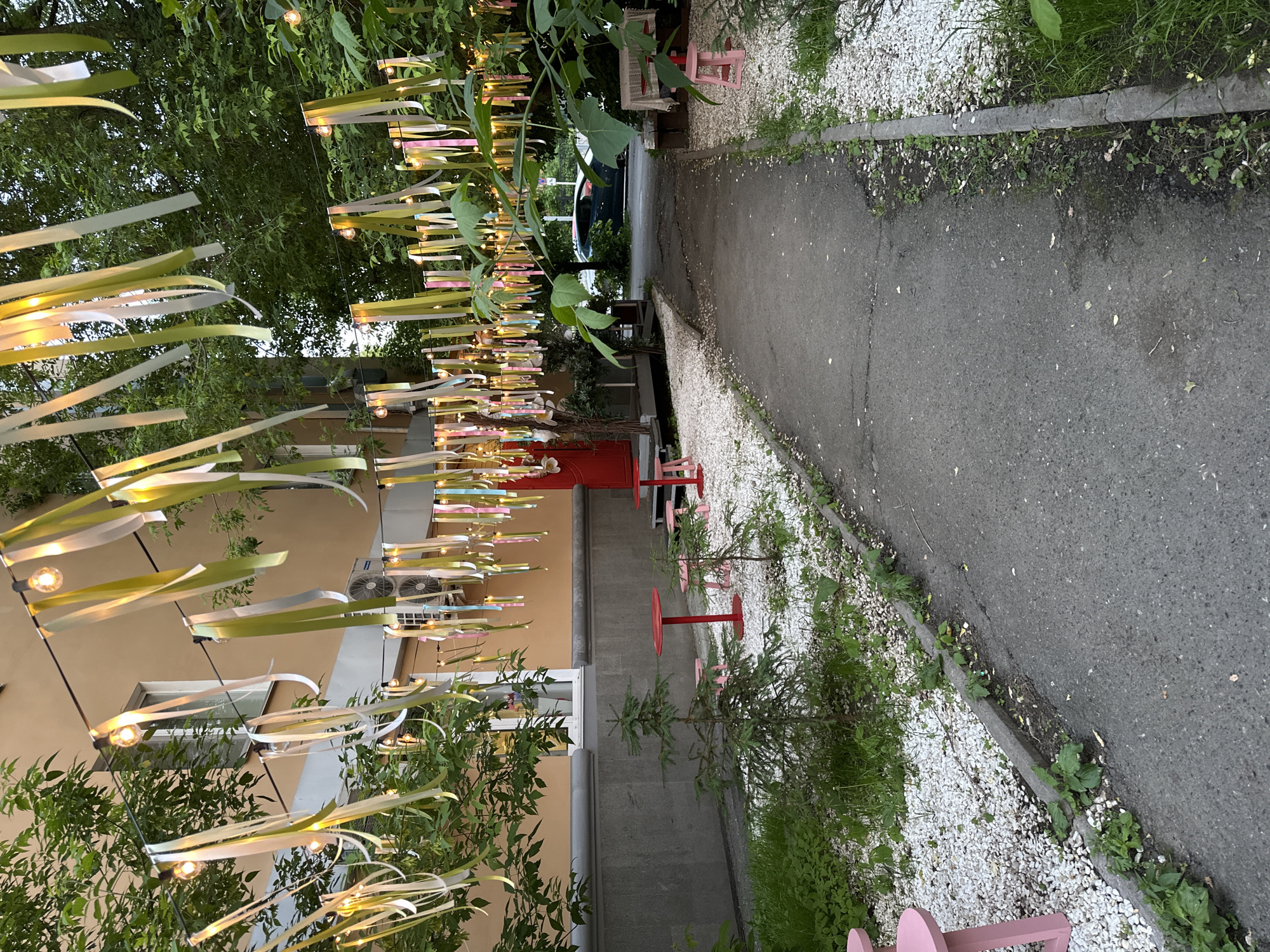 This image presents a decorated outdoor alleyway or courtyard in Perm, Russia, designed for leisure or dining. The scene is characterized by a festive ambiance created by overhead strings of lights and numerous ribbons. These ribbons, in shades of pale green, white, and subtle pink or purple, are strung across the space, possibly between trees or structures, forming a whimsical canopy.
The ground features an asphalt path running vertically along the right side of the frame, bordered by a strip of white gravel interspersed with small green plants and weeds. A concrete curb separates the path from the gravel area. On the left, a light beige building facade is visible, featuring a window and an outdoor air conditioning unit. A vibrant red door stands out against the building wall further back.
Throughout the area, natural elements are prominent, including lush green foliage from trees and bushes overhead and along the left, providing a natural backdrop and canopy. Smaller, coniferous-looking plants are also placed in the gravel strips. Outdoor furniture includes several small, minimalist tables with red tops and matching pink chairs, suggesting an inviting seating arrangement. A dark-colored car is partially visible in the background, beyond the main decorated area.
The glowing lights indicate that the photograph was taken during dusk or early evening, contributing to a warm and inviting atmosphere. No people are present, lending a tranquil mood to the whimsical setting. There is no visible text in the image.