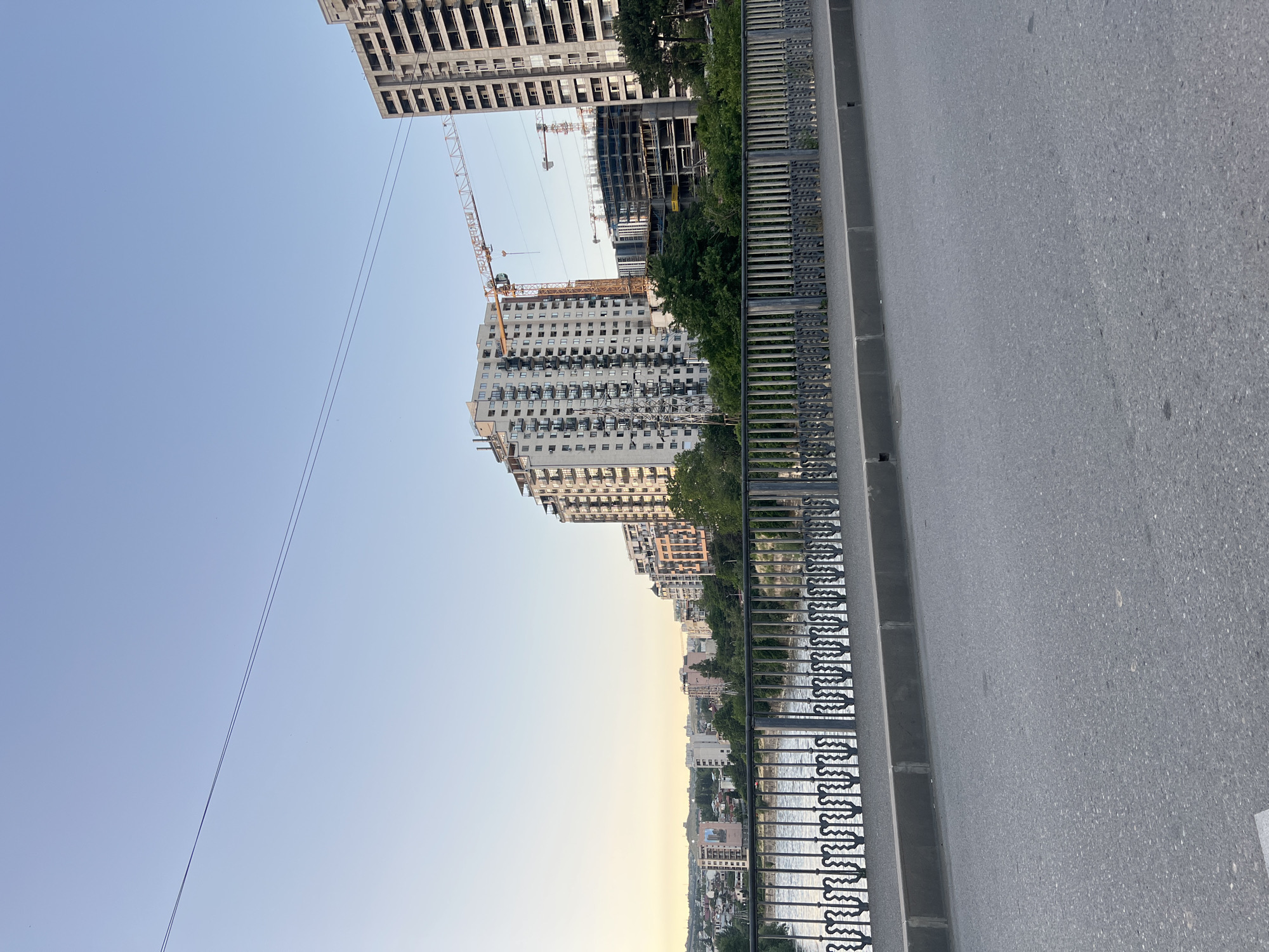 This image depicts a cityscape with a river and multiple buildings, some of which are under construction. In the foreground, a road runs parallel to a decorative metal fence. Beyond the fence, a river flows, and on the opposite bank, a line of residential buildings is visible. Further in the background, several taller apartment buildings are prominent, with construction cranes suggesting ongoing development. The sky is clear with a light gradient of color, indicating it might be late afternoon or early evening. The scene appears to be in Tbilisi, Georgia, given the characteristic architecture and the presence of the Mtkvari River. There are no people visible in the frame.