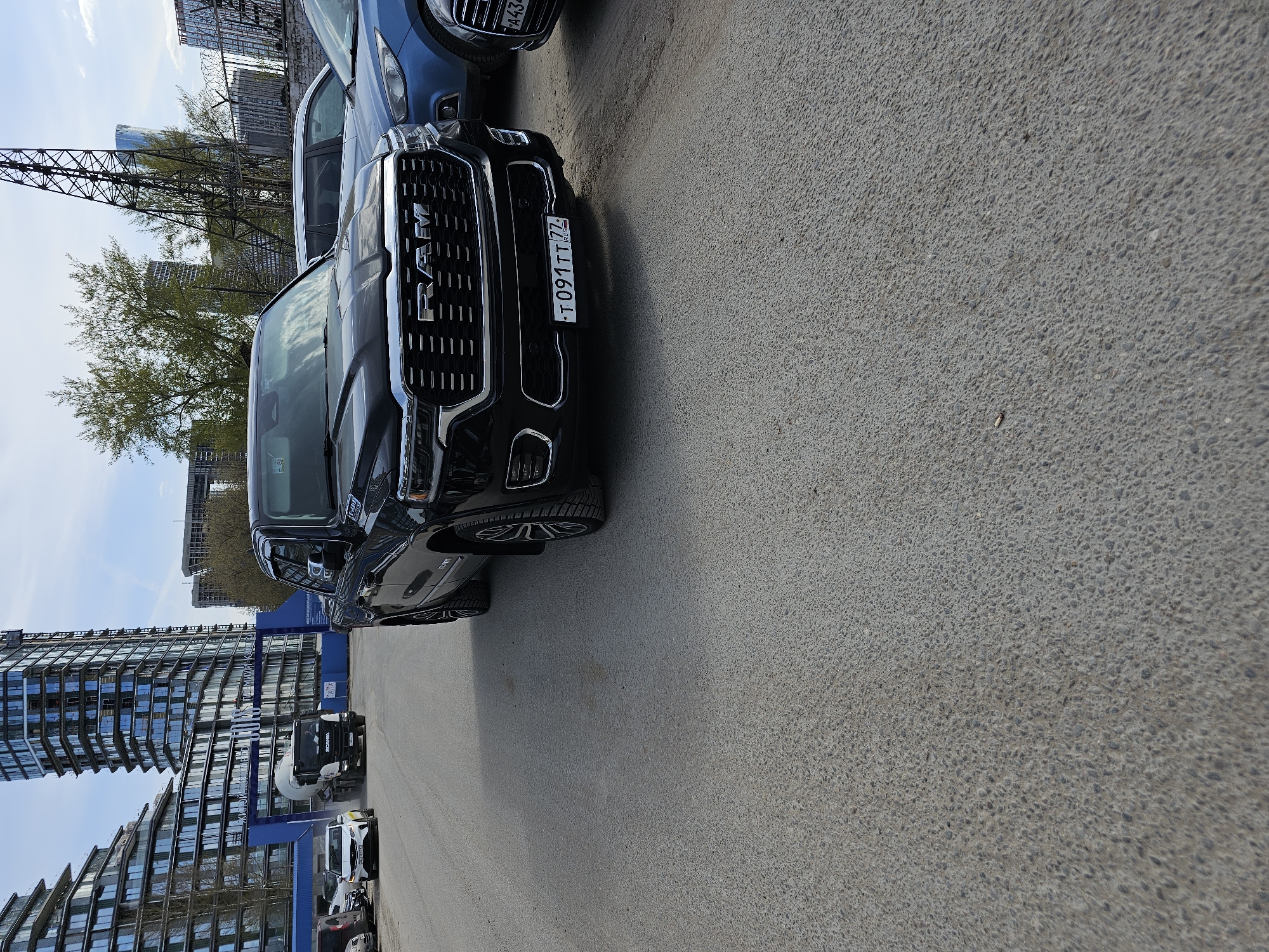 The image shows a parked black RAM truck with a Russian license plate "T 091 TT 77" on an asphalt parking lot. The truck is positioned at an angle to the viewer. Behind the truck, there is a modern building with a glass facade and a unique architectural design. A construction crane is visible in the background along with some trees. In the distance, there are other vehicles, including a white van and a cement truck. The scene appears to be located in a modern urban area, likely in Moscow, Russia, given the license plate information. The weather is sunny, and the time of day appears to be daytime.