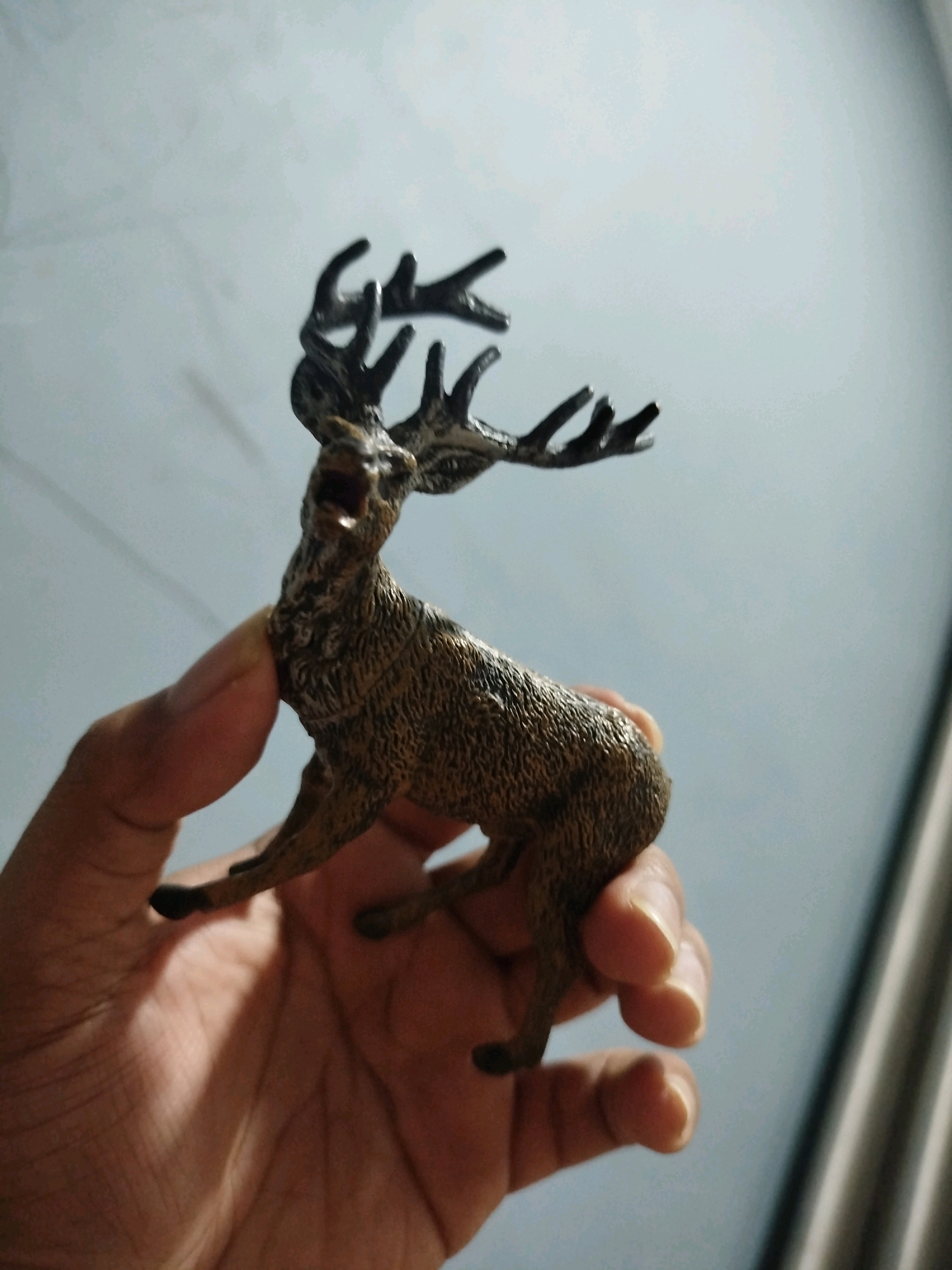 A person's hand is holding a miniature toy deer. The deer is brown and has antlers. The background is a light blue wall and a gray curtain. The image was taken in Mojoagung, Indonesia.