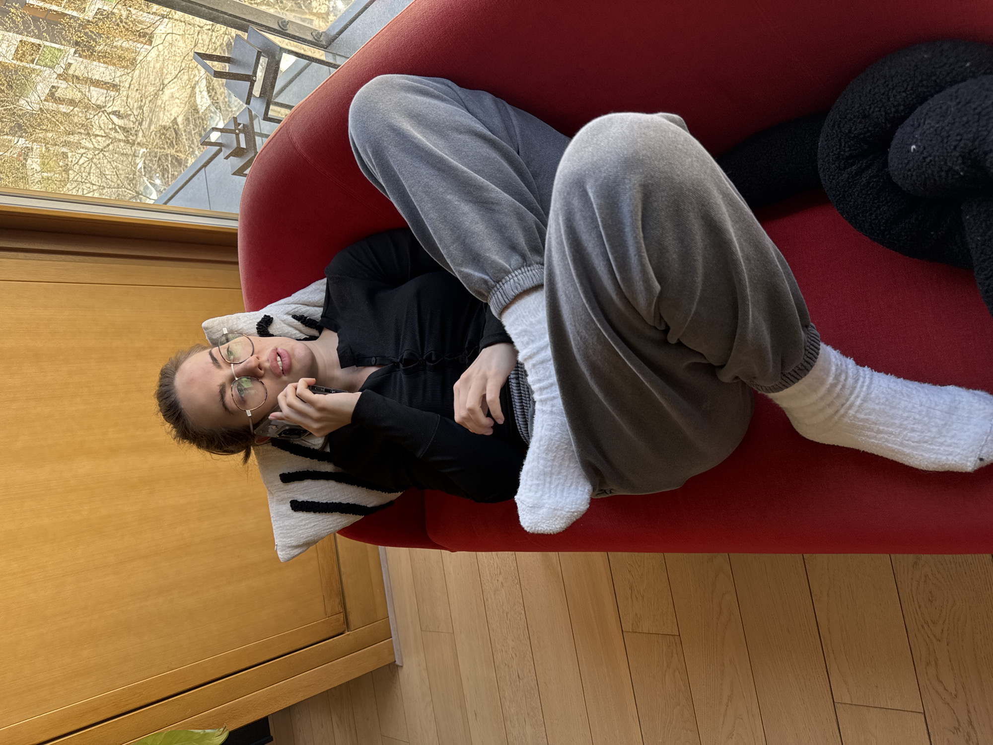 A young woman is depicted lying on her back on a vibrant red sofa, looking towards the camera with a neutral expression. She is wearing grey sweatpants, a black long-sleeved top, and white socks. Her eyeglasses are perched on her nose. Her head rests on a white pillow adorned with black horizontal stripes. She holds a silver-colored smartphone to her left ear, suggesting she might be on a call or interacting with the device.
The setting appears to be indoors, possibly a living room or lounge area in Warsaw, Poland, beside a large window that reveals an outdoor scene. Through the window, bare tree branches, possibly signaling late winter or early spring, are visible under bright daylight. A modern architectural structure, possibly part of a building or balcony, can also be seen outside. Below the window, there is a warm-toned wooden cabinet or wall panel. The flooring in the room is made of light-colored wooden planks. Behind the woman's legs, on the sofa, a black, textured, knot-shaped object or pillow is partially visible. The image is captured from a slightly elevated perspective, giving a clear view of the woman and her immediate surroundings. No visible text is present in the image.