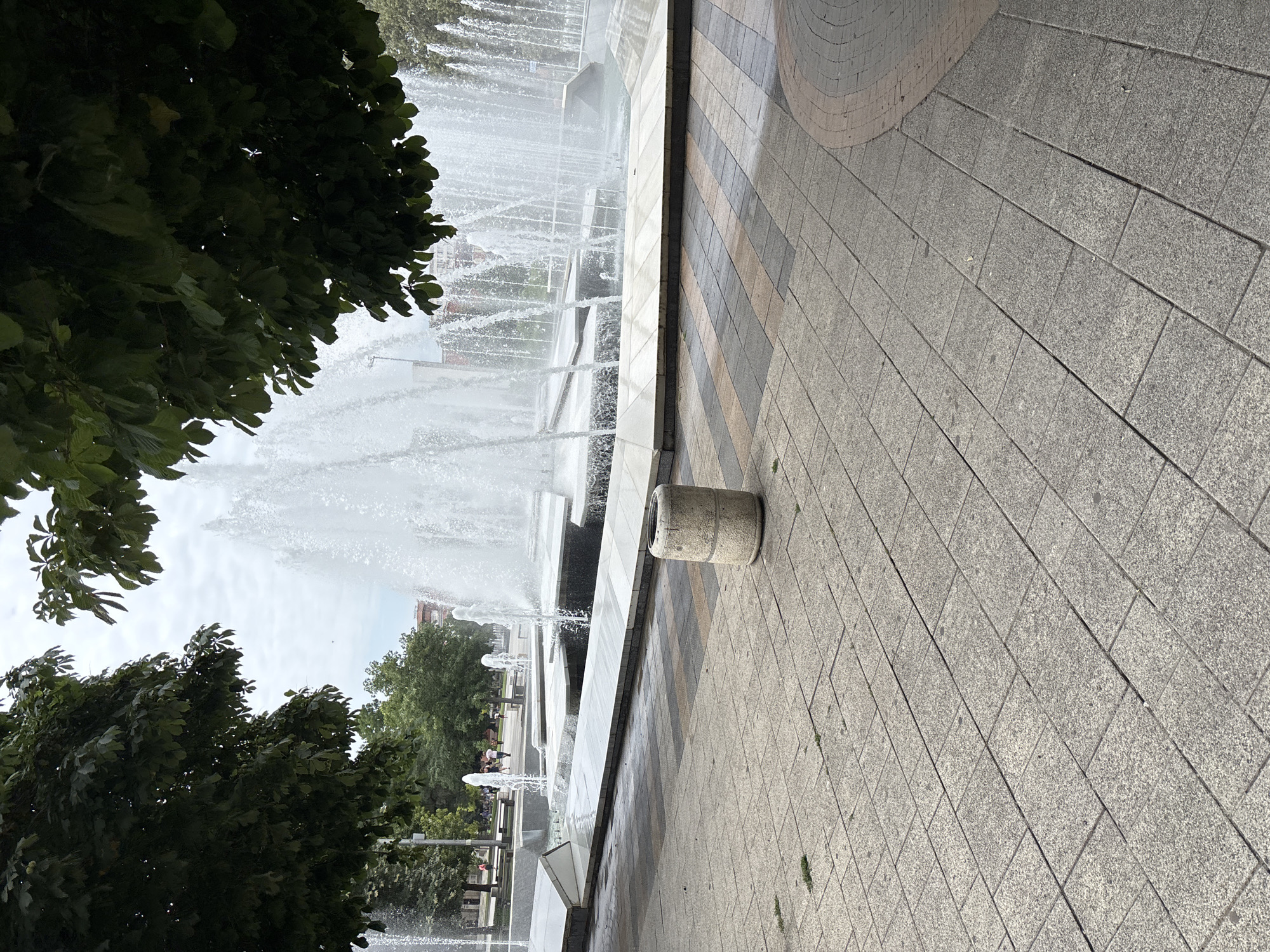 The image shows a public fountain with multiple jets of water spraying upwards. The fountain is made of concrete and stone and is surrounded by a paved area. Trees with green leaves frame the top of the image, and a cloudy sky is visible in the background. In the foreground, there is a cylindrical concrete trash bin. People are visible in the background, walking in a park-like setting. The scene appears to be a public space in a city, likely a park or plaza. The image is captured from a low angle, looking up towards the fountain and trees. The location is identified as the center of the city of Pleven, Bulgaria, specifically the "Fountain of the Youth" or "Fontan" in the city's main pedestrian street.