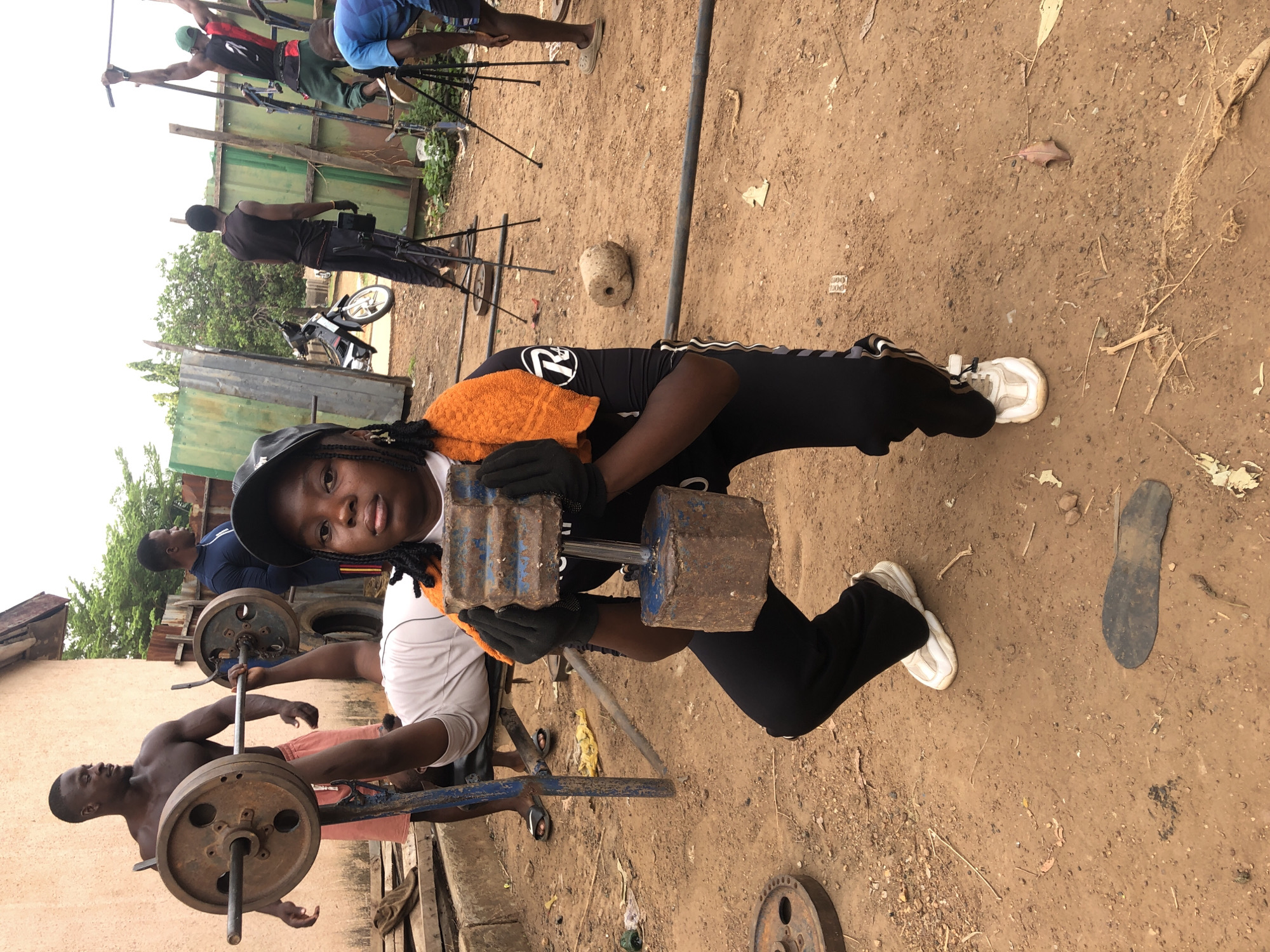 This media file captures an outdoor scene in Akwanga, Nigeria, showing a makeshift gym environment, possibly serving as a film set given the boom mic and other personnel.
The central figure is a young person, likely female, with dark skin and braided hair peeking from under a black cap. She wears a white long-sleeve shirt with an orange towel draped over her shoulder, black athletic pants, black gloves, and white sneakers. She is squatting slightly, looking directly at the camera with a neutral expression, and holding a heavy, rustic-looking dumbbell. The dumbbell features two rectangular, blue-painted, and visibly rusty weights on a silver bar. The number "72" is prominently displayed on her black top.
Around her, several other individuals are present. In the immediate foreground to the left, a shirtless man with dark skin is engaged in weightlifting, pushing up a barbell with large, dark, circular weights. Further back, another man in a dark blue shirt observes. Towards the back-left, a man in a black tank top and pants stands beside a small motorcycle. In the background, to the far left, two men appear to be part of a production crew: one in a red and black shirt holds a boom mic on a stand, and another in blue shorts is partially visible.
The setting is an unpaved area with light brown, dusty dirt ground. Various pieces of old or handmade weightlifting equipment are scattered around, including additional barbell weights and metal stands. The background features a simple, ad-hoc fence made of corrugated metal sheets and wooden planks, with some greenery visible over the top. The sky is bright but appears overcast, indicating daytime. A small, light-colored object on the ground bears faint numbers like "200" or "300." The overall scene suggests a vibrant, active atmosphere in a practical, outdoor training or filming space.
