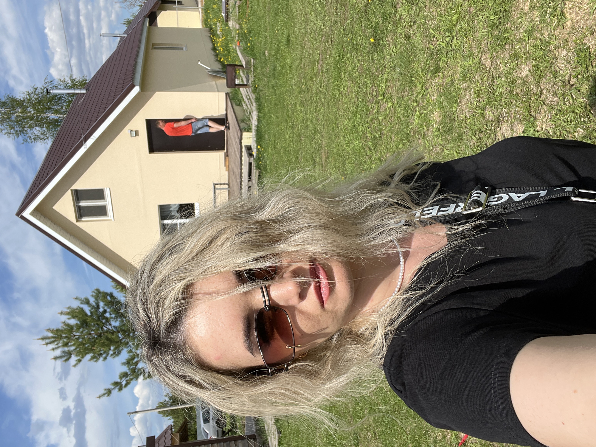 The image is a selfie of a woman in Гаревая, Russia. She is fair-skinned with blonde, wavy hair and is wearing sunglasses, a black shirt, and a pearl necklace. She has a "Karl Lagerfeld" strap on her person. In the background, there is a one-story house with a brown roof and a light yellow exterior. A person in a red shirt and blue shorts stands in the doorway of the house. Green grass covers the ground. The sky is blue with scattered clouds. A tree is visible to the left of the house.