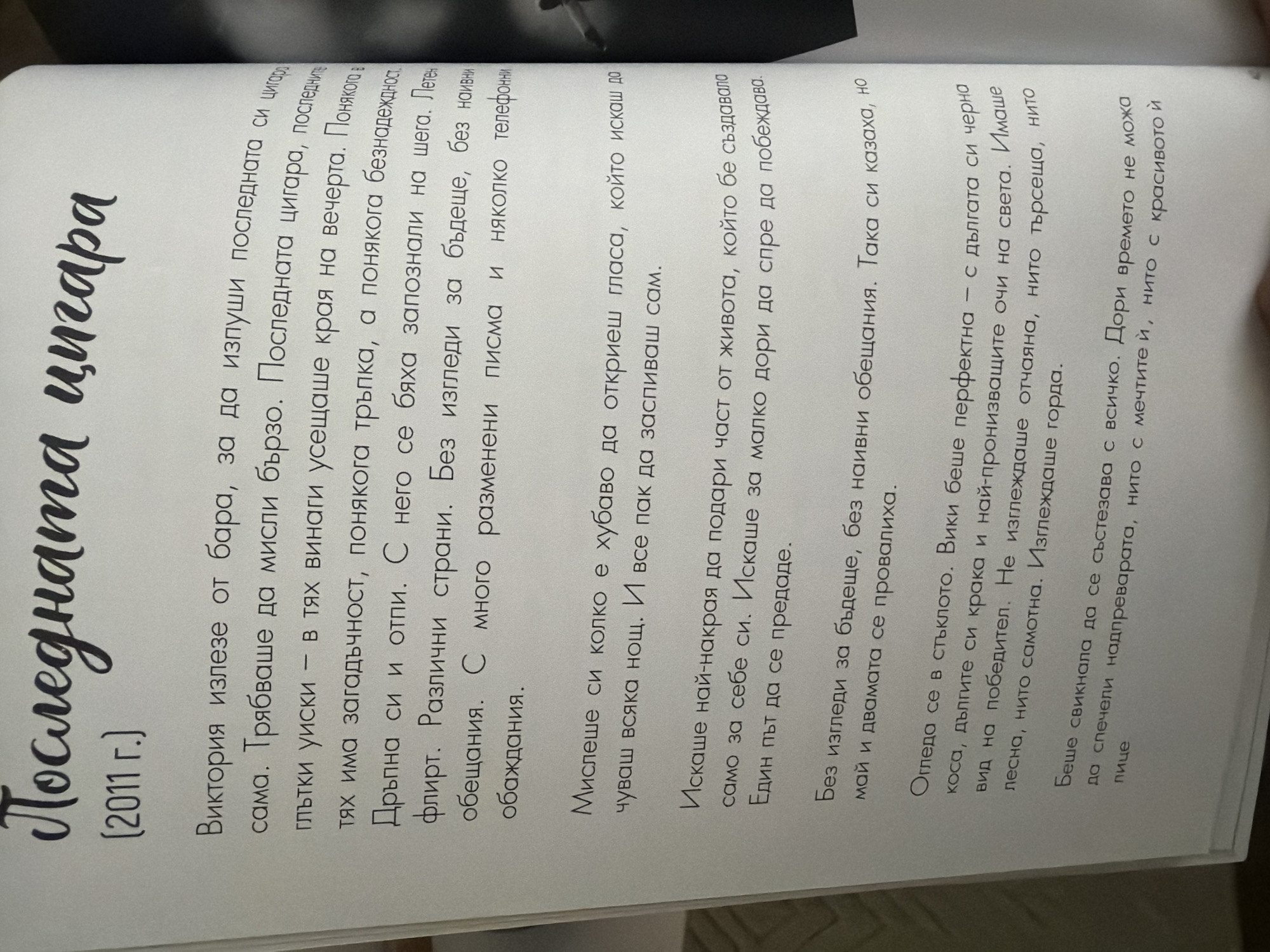 The image shows a close-up of an open book with Bulgarian text. The book is lying flat, and the pages are slightly curved. The text is printed in a dark font, and there are some handwritten annotations or markings visible in some areas. The title of the book appears to be "Последната цигара" (The Last Cigarette), with the year "(2011 г.)" below it. The content of the book is narrative prose.
The image itself does not contain any visual elements that would indicate its location in the city of Pazardzhik, Bulgaria. The scene is a still life of a book, with no contextual clues about its geographical origin.