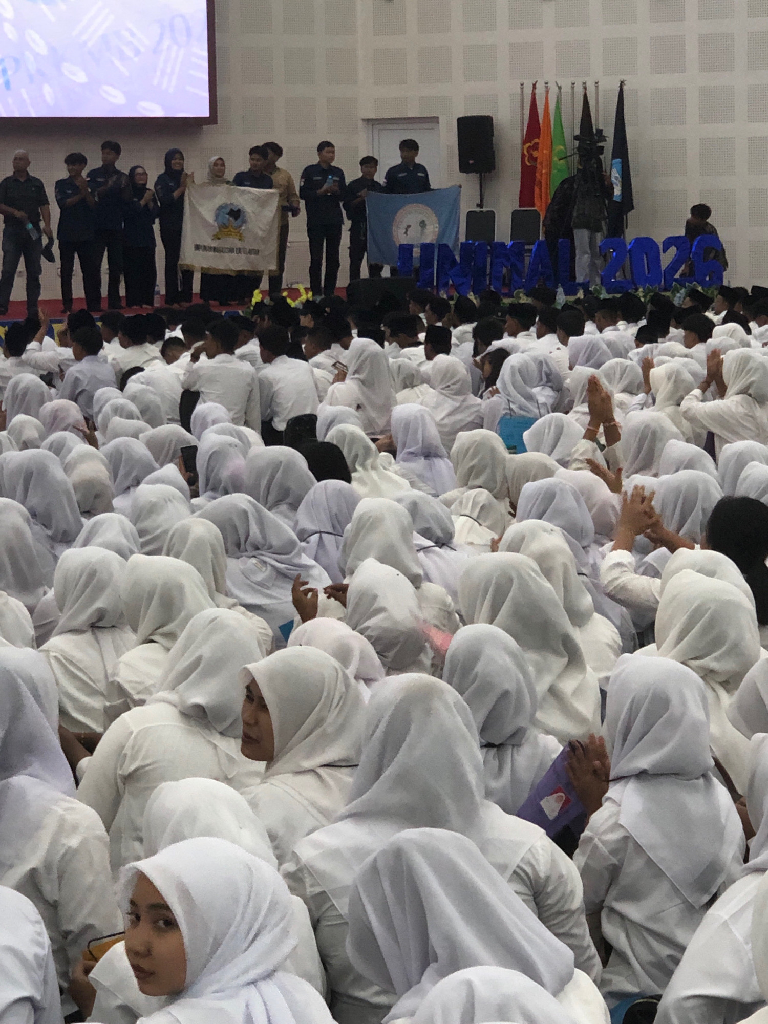 A group of people, predominantly young individuals wearing white Islamic attire, are gathered in an indoor space. Many are seated, facing a stage area where several adults stand holding flags and a banner. Behind the stage, large blue block letters spell out "JIMNAL 2026," suggesting an event or organization associated with that year. The room appears to be a hall or auditorium with a tiled wall and a large screen at the back. The overall atmosphere seems formal and possibly celebratory or commemorative, given the presence of flags and banners. The location is identified as Reuleut Timu, Indonesia.