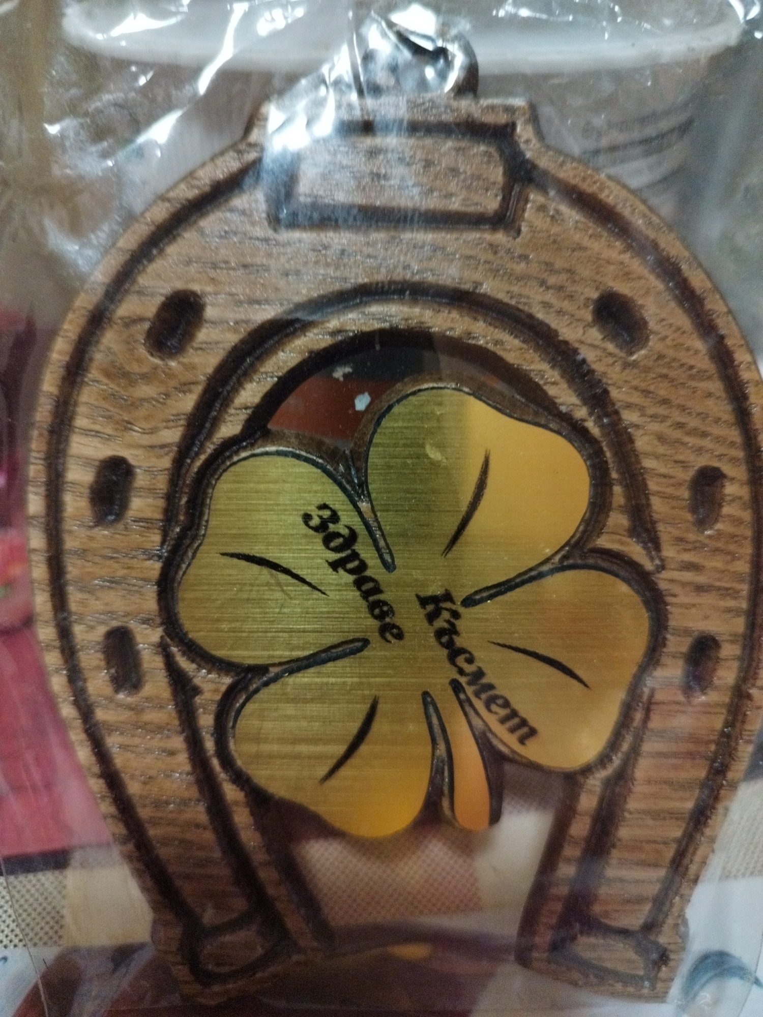 The image shows a wooden horseshoe-shaped souvenir with a golden four-leaf clover at its center. The words "Здраве" (Health) and "Късмет" (Luck) are written on the clover in Cyrillic script. The horseshoe has several holes, likely for decorative purposes. The souvenir appears to be packaged in clear plastic. The location is inferred to be Sofia, Bulgaria, based on the Cyrillic script and the type of souvenir.