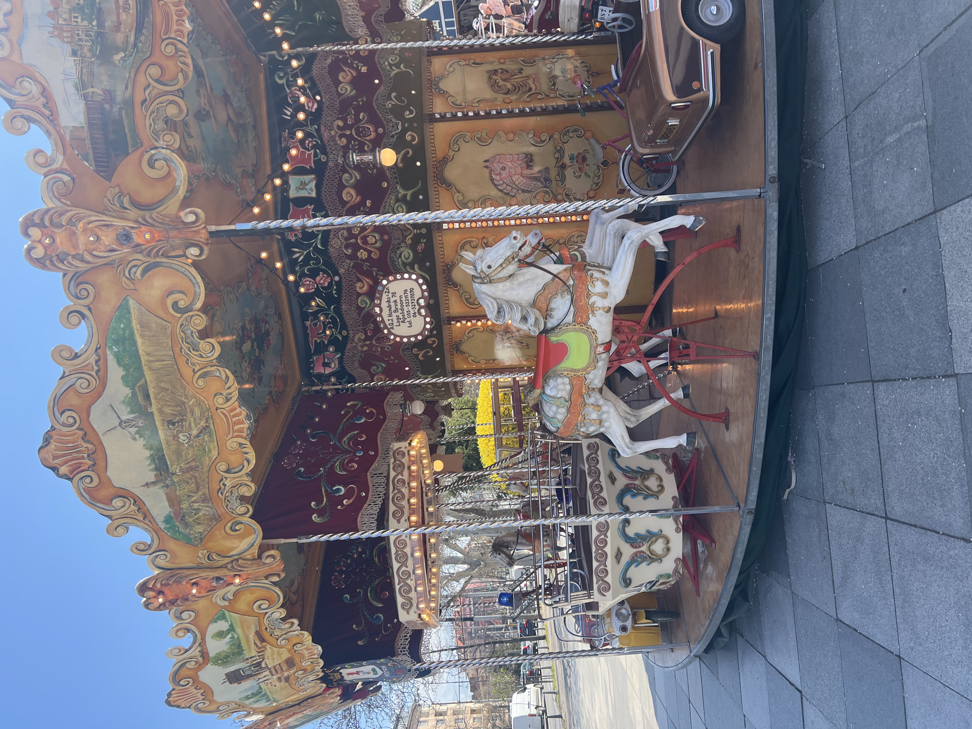 The image shows a carousel in Sofia, Bulgaria. The carousel features ornate decorations, including painted horses and other figures, with string lights illuminating the structure. The carousel is located on a cobblestone paved area, possibly a park or plaza. In the background, city buildings and trees are visible, suggesting an urban setting. The sky is clear and blue.