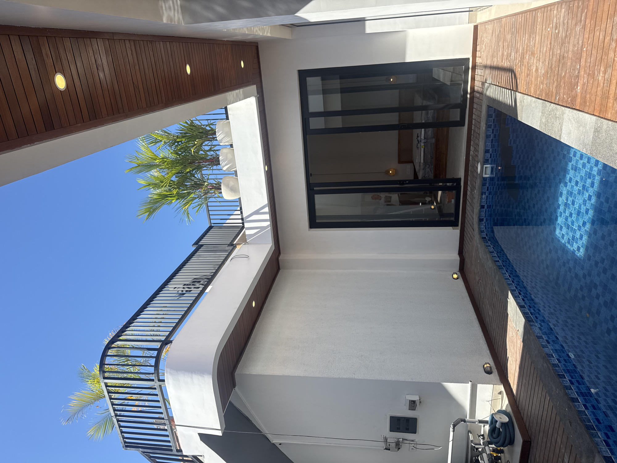 The image captures a modern architectural design, likely a private residence with a pool, in Denpasar, Indonesia. The design features a two-story structure with a balcony enclosed by black metal railings on the upper level. Tropical plants, including palms, are visible on the balcony. The building’s exterior is predominantly white, with sections of wooden paneling. A sliding glass door with a black frame provides access to an interior space. Adjacent to the building is a pool with blue mosaic tiles, bordered by wooden decking and concrete. Below the pool, there are the filtering and pumping infrastructure and electrical outlets. The clear blue sky indicates a sunny day.