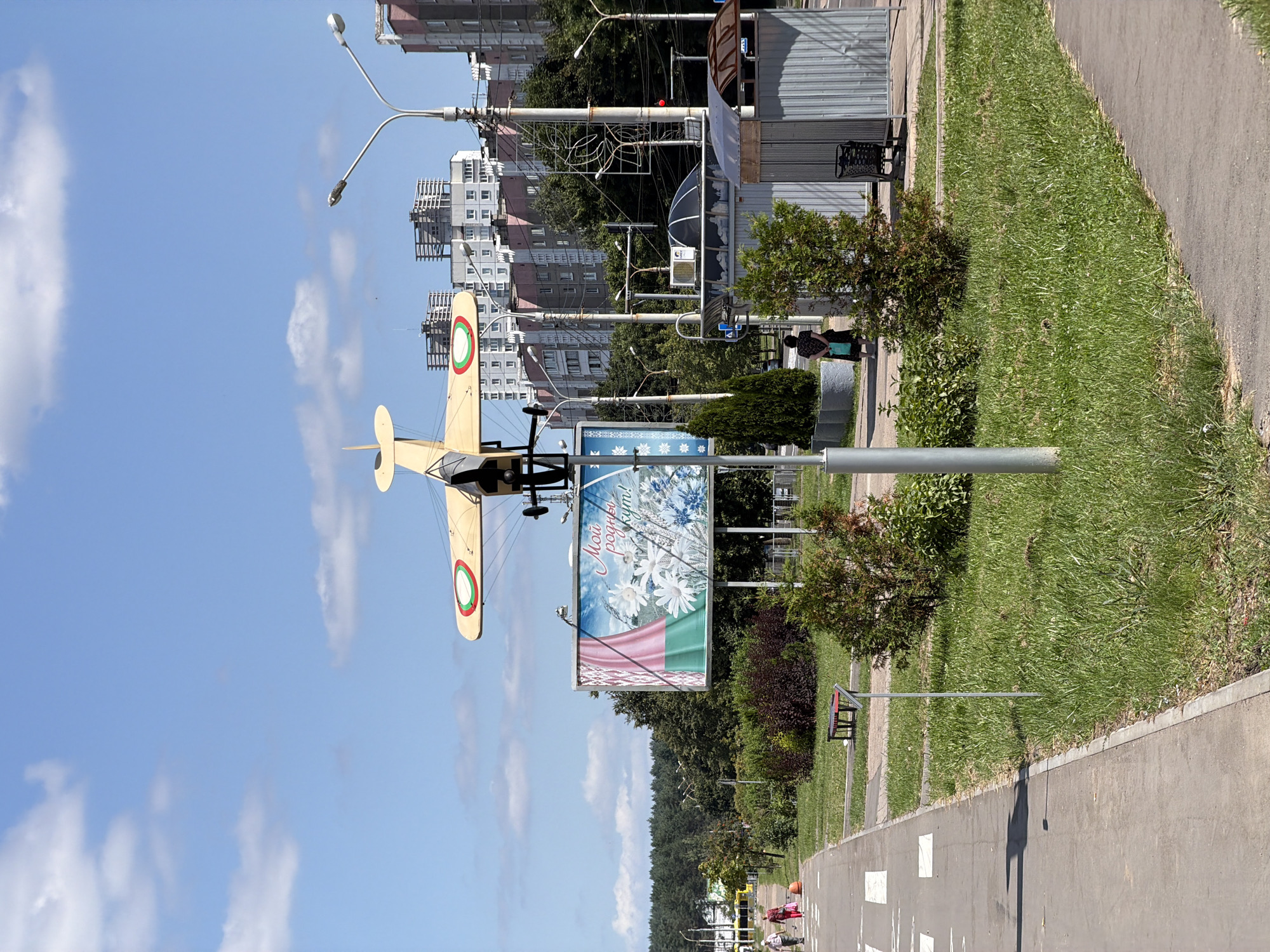 The image depicts a public space in Minsk, Belarus, with an iconic biplane sculpture mounted on a pole. The biplane is yellow with red and green circular markings on its wings. In the background, several multi-story apartment buildings are visible, some under construction. To the left of the biplane sculpture is a large billboard featuring the Belarusian flag colors and text in Cyrillic script, reading "Мои родны куты!" (My native corners!). A paved walkway runs along the left side of the image, with two women walking away from the camera. Green grass and small trees are present in the foreground and middle ground. Streetlights and what appear to be bus stops or small utility structures are also scattered throughout the scene. The sky is blue with scattered white clouds.