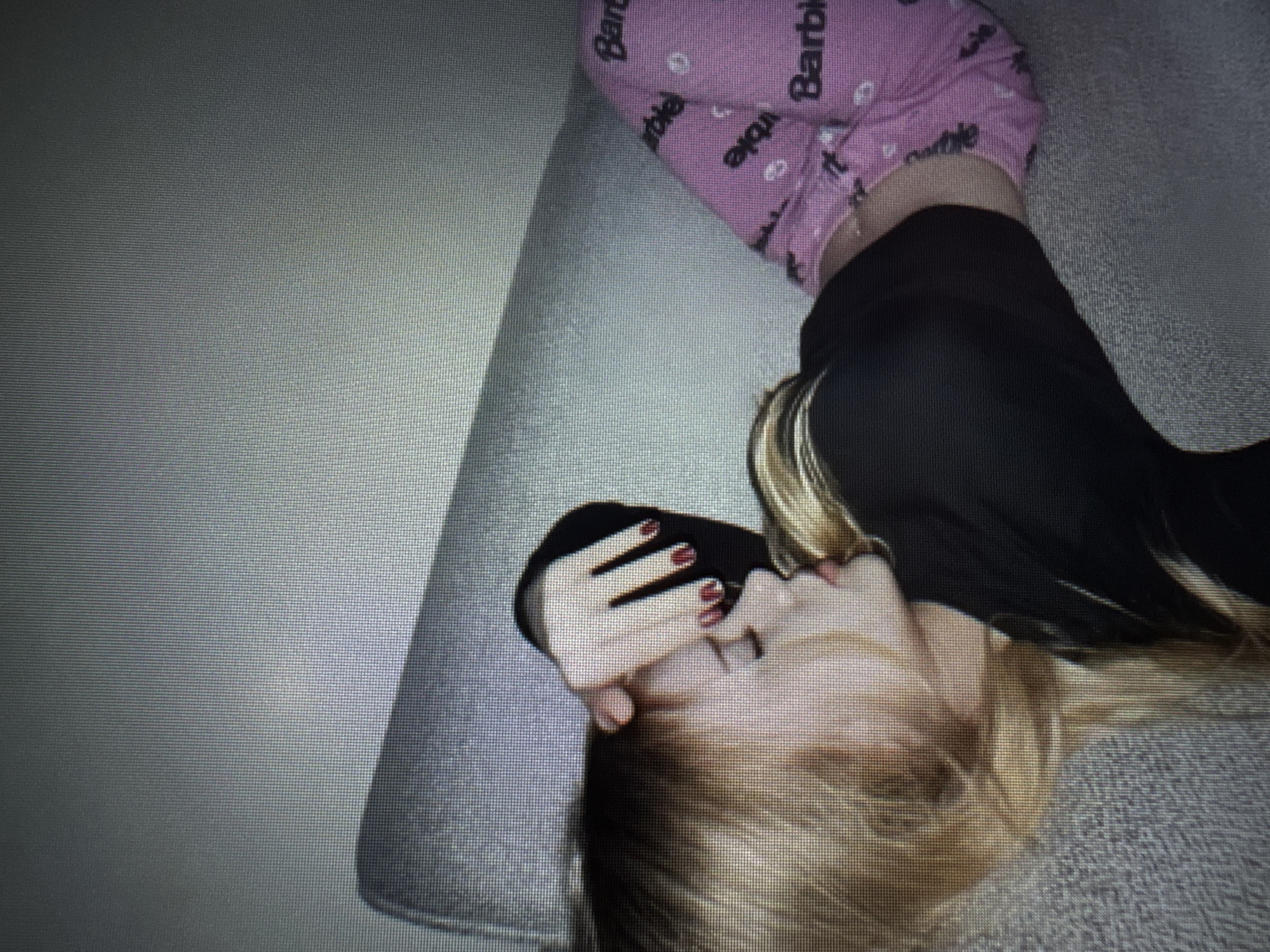 The image displays an angled, close-up view of a person lying down indoors, likely on a grey, textured surface such as a sofa or carpet. The perspective is rotated, showing the person's head towards the bottom right and legs extending towards the top left. The subject has blonde hair and is wearing pink pajama-style pants repeatedly printed with the word "Barbie" and small white circular patterns. They are also wearing a black top or shirt. One of the person's hands is raised near their face, possibly touching their chin or mouth, and features bright red nail polish. The facial expression is not fully discernible due to the angle and partial obstruction. The entire image is characterized by a distinct pixelated or grid-like pattern, suggesting it might be a photograph of a digital screen or processed to achieve a similar effect. The setting appears to be a casual indoor environment, indicative of rest or relaxation.