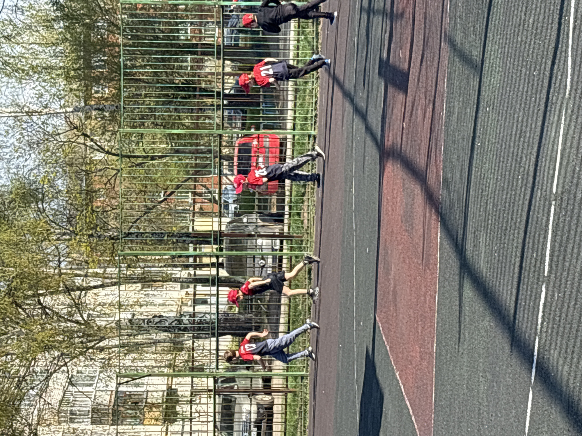A photograph shows several children running on an outdoor sports court with a green fence in Izhevsk, Russia. The court is divided into maroon and dark gray lanes with white lines. Five children are visible, all wearing red hats and sports jerseys. Some are wearing black shorts, and others are wearing gray pants. They appear to be running in a line. Behind the fence, there is foliage and a building visible, along with what appears to be a red vehicle.