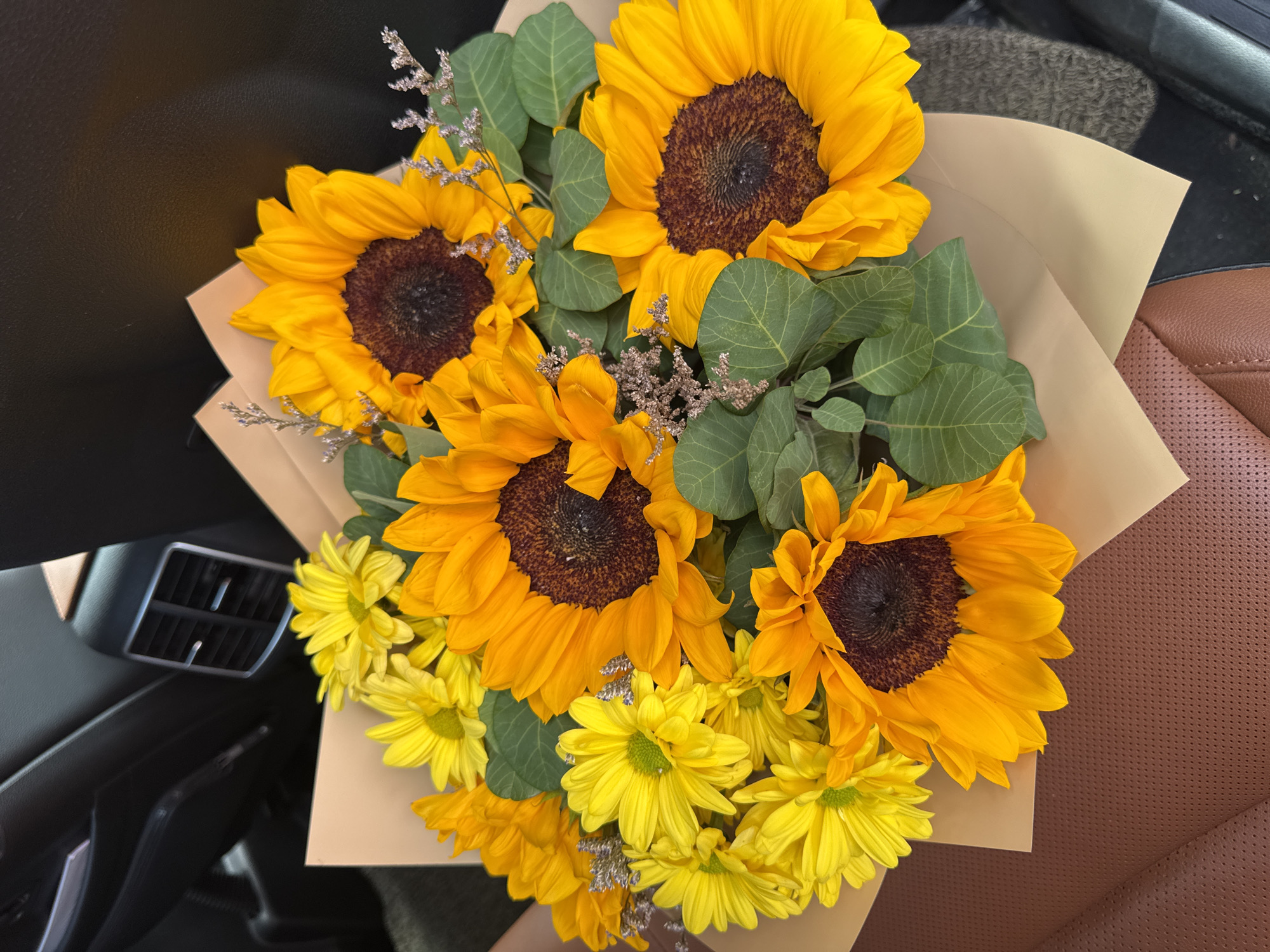 A bouquet of sunflowers and yellow daisies is presented on a background of car interior. The bouquet is wrapped in brown paper. The sunflowers are large with dark brown centers and bright yellow petals. The yellow daisies are smaller and have light green centers. Green leaves and some dried filler flowers are interspersed throughout the bouquet. The background shows the textured brown leather of a car seat, a dark car interior with ventilation grilles, and a grey carpet. The scene is likely captured in Almaty, Kazakhstan, given the context.