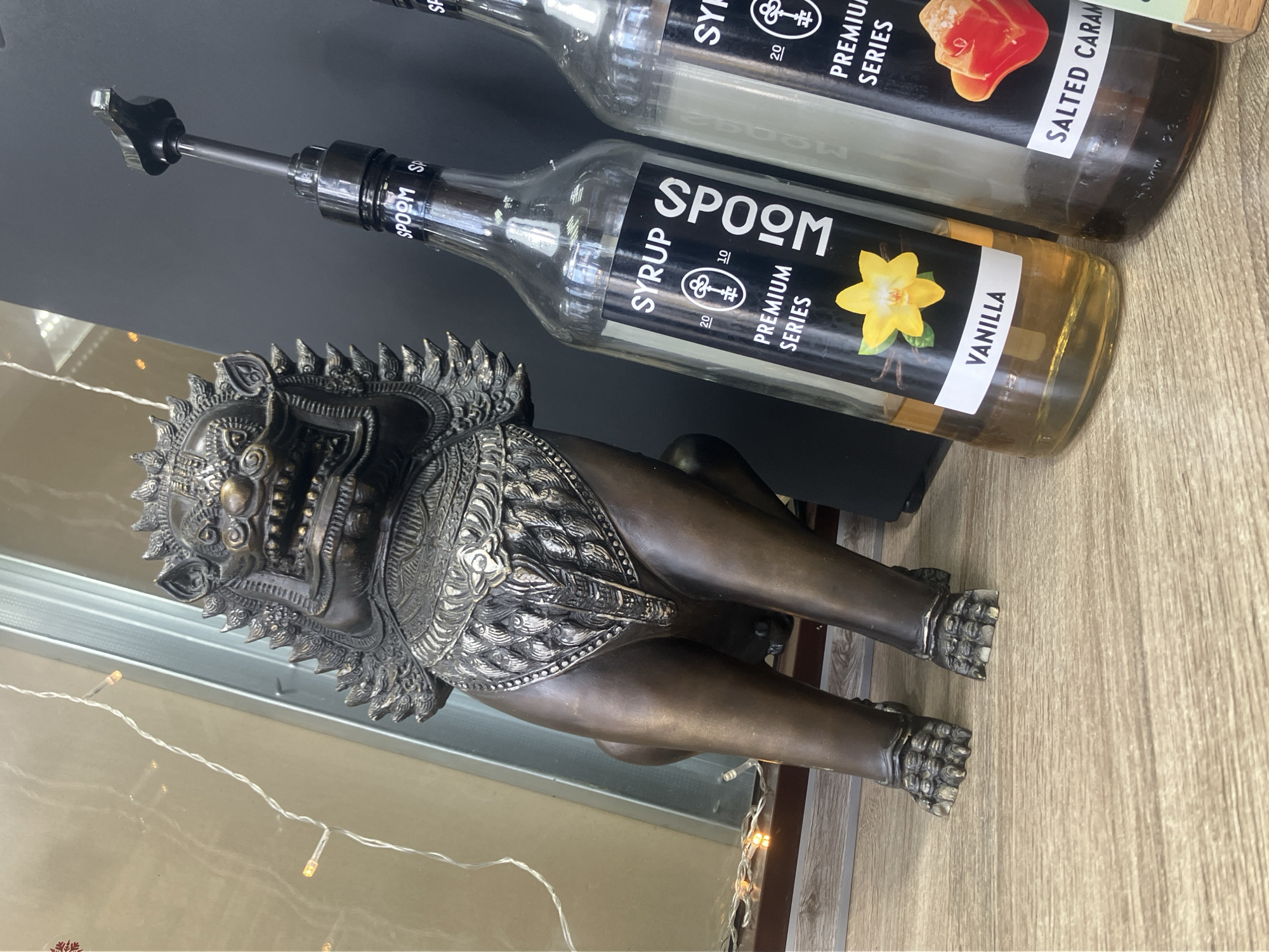 This image appears to be taken indoors, possibly in a cafe or shop setting. The scene includes two bottles of syrup from a brand called "SPOOM." One is Vanilla-flavored, and the other is Salted Caramel. A decorative, metallic statue of a mythical lion-like creature rests nearby. The background appears to be a mix of reflective glass and dark, solid surfaces, along with a wood-grain-patterned countertop. The city of Chaikovsky, Russia, cannot be determined from the visual elements present in the image.