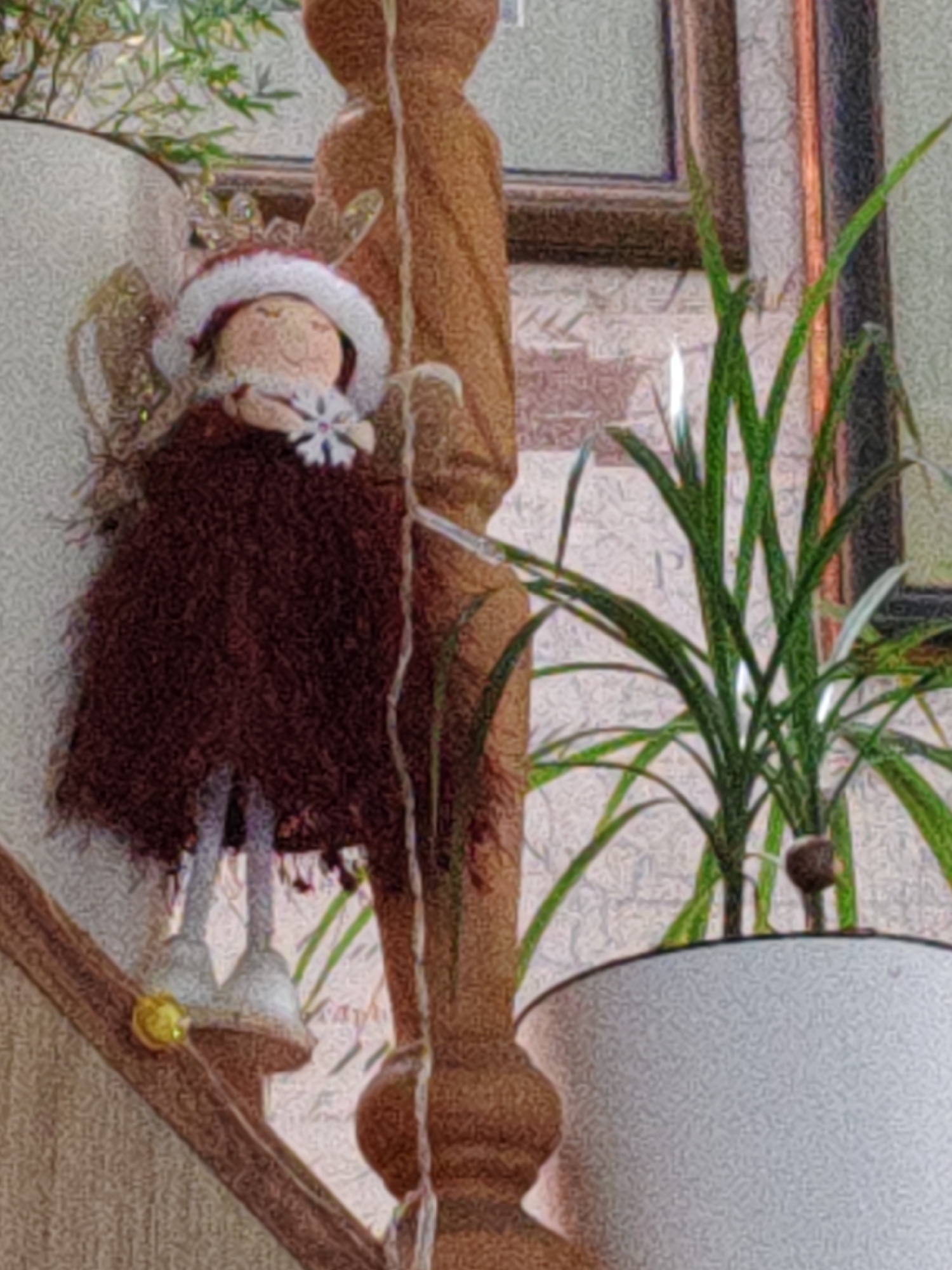 The image shows a close-up of a decorative doll with reindeer antlers and a fluffy red dress, wearing a white hat trimmed with red. The doll is holding a snowflake ornament. The doll is positioned on a wooden surface that appears to be part of a staircase or railing. In the background, a green plant in a white pot is visible, along with a wooden pillar and a framed picture. The overall scene suggests a festive or holiday decoration, likely indoors. There are no discernible elements that would indicate a specific location within the city of Rostov-on-Don, Russia.