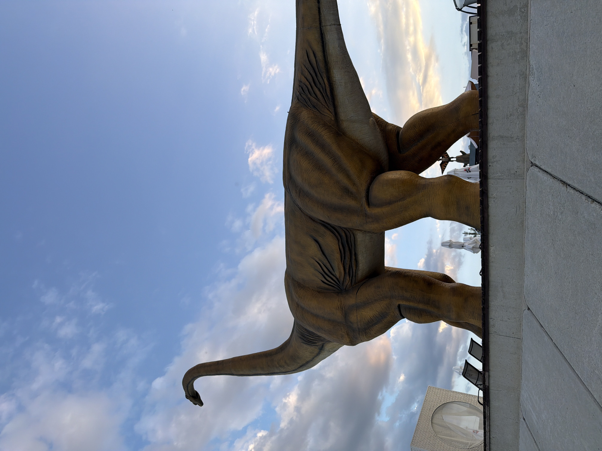 A large, tan, dinosaur statue with visible muscle definition and patterned skin is positioned prominently in the foreground, facing left. Its long neck curves upwards and to the left against a backdrop of a cloudy blue sky. The dinosaur stands on a raised platform, a concrete structure that extends horizontally across the lower portion of the image. Behind the dinosaur, on the platform, are several lounge chairs, some white umbrellas, and a modern, white, cube-shaped structure with a circular opening. The scene suggests an outdoor recreational area, potentially a resort or park. The location is identified as Fontanka, Ukraine.