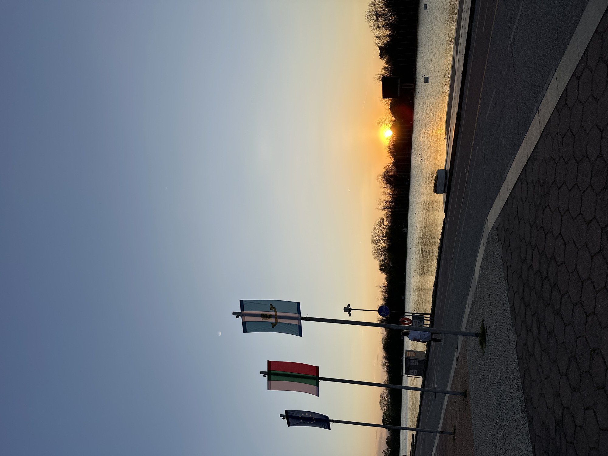 The image shows a sunset scene in Plovdiv, Bulgaria, captured from a slightly elevated perspective. The sun is setting over a body of water, possibly a lake or river, reflecting light on the water's surface. Silhouetted trees and vegetation line the opposite bank.
In the foreground, there are three flags displayed on poles. From top to bottom, they appear to be the flag of Plovdiv, a red and green horizontal striped flag (possibly the flag of Transnistria), and the flag of the European Union. A sidewalk and road run along the water's edge, adjacent to the flag poles. A person stands nearby, partially obscured.