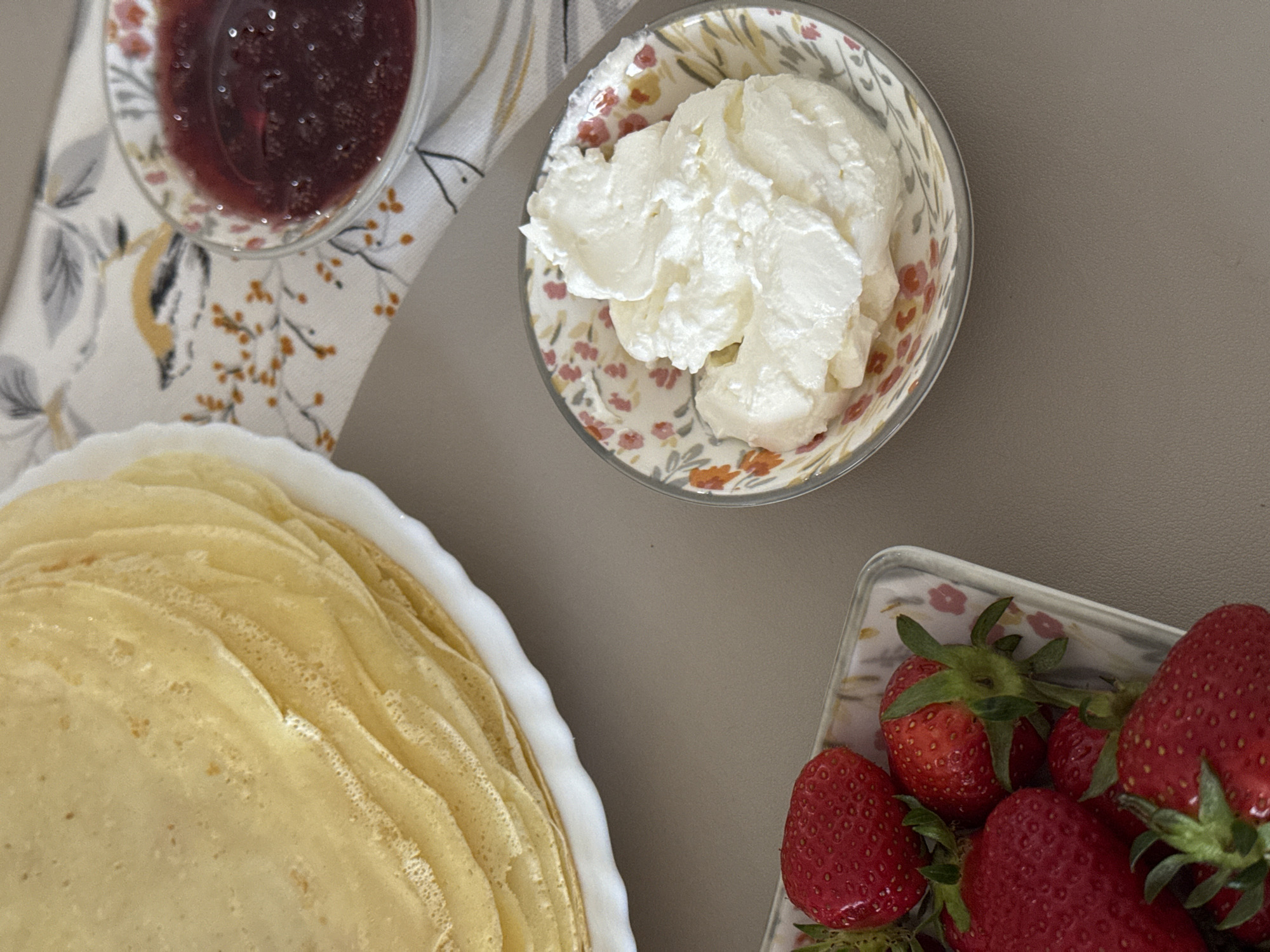The image shows a tabletop scene with various food items arranged for consumption. In the lower-left, a white dish holds a stack of crepes. Above the crepes and slightly to the right is a floral-patterned bowl filled with a dollop of white cream. A clear glass dish filled with red jam is situated above the crepes and to the left of the cream. In the lower-right, a clear rectangular dish contains several strawberries with green stems. The background surface appears to be a neutral, solid color. A floral-patterned napkin is partially visible near the jam. The location is not discernible from the image.