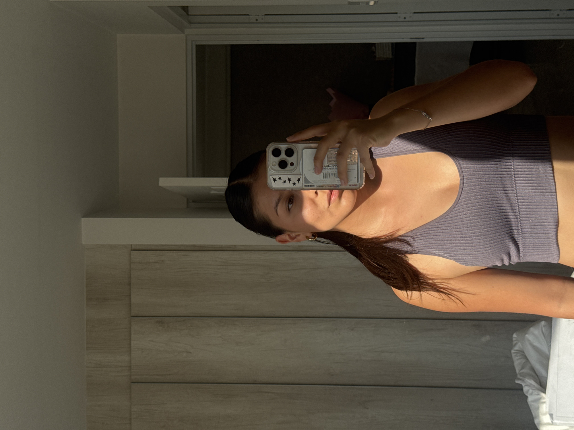 A young woman is captured in a mirror selfie, held by her right hand. She has dark hair styled in a sleek ponytail and is wearing a ribbed, light purple or grey crop top. Her expression is neutral as she looks into the phone's camera. Strong, direct sunlight illuminates the right side of her face, arm, and torso, creating sharp highlights and shadows. A delicate silver-colored bracelet adorns her right wrist, and a small gold hoop earring is visible on her left ear.
She holds an iPhone in a clear case, which features several black star stickers on the bottom left corner. Inside the phone case, behind the phone itself, a piece of paper or card is visible with faint, reversed text including "adlibu ww," a barcode-like pattern, and the date "01/10/20."
The setting is indoors, with a wooden dresser or cabinet forming part of the background to the left. A white shelf or surface runs above the dresser. The reflection in the mirror shows a darker room or area behind the photographer, with a hint of a window frame and a soft pink object dimly visible. The prominent natural light suggests it is daytime. The image provides no specific visual cues for San Francisco Ocotlán (Ocotlán), Mexico, beyond the provided context.