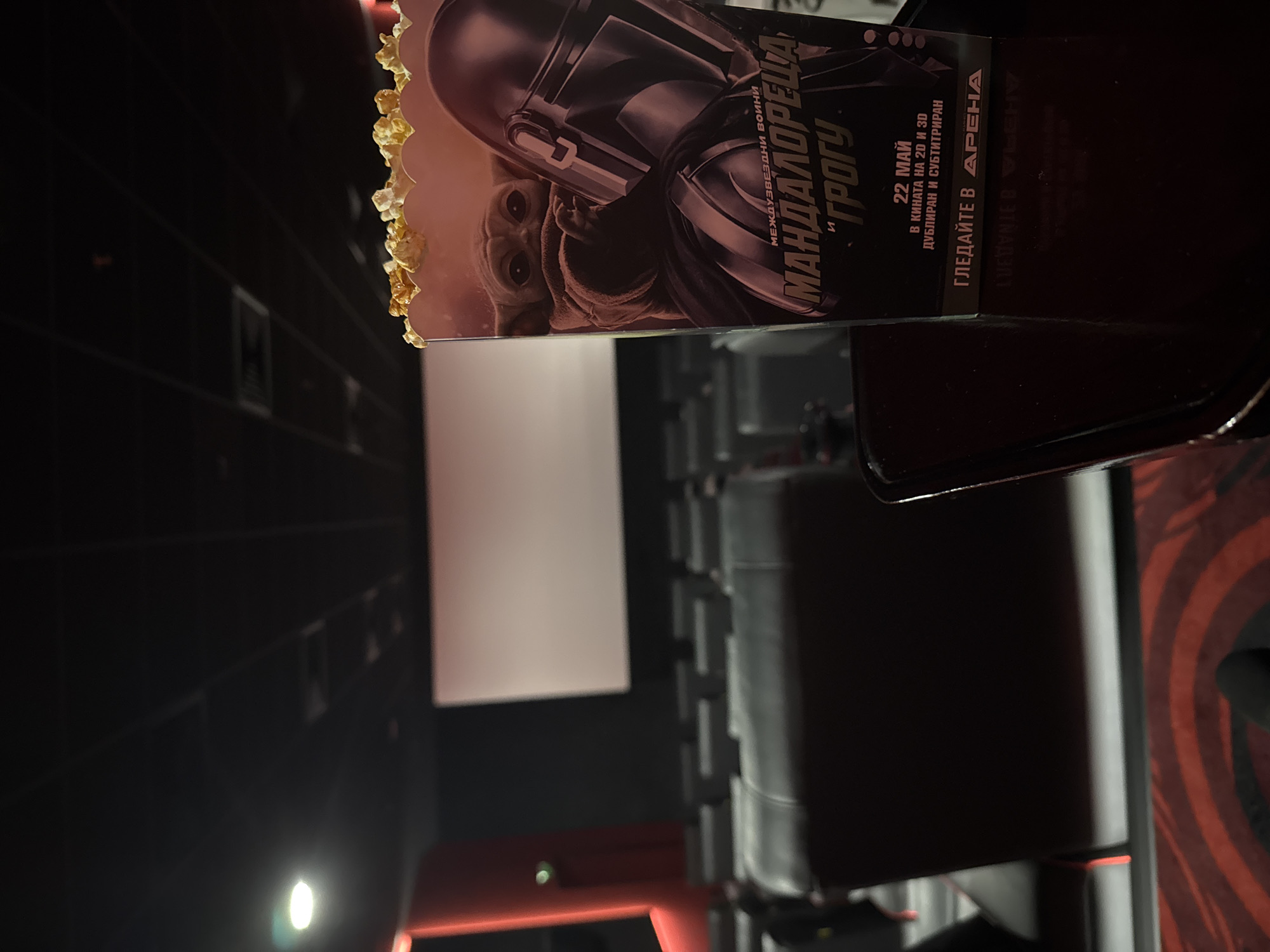 The image shows a movie theatre in Sofia, Bulgaria. A box of popcorn with The Mandalorian and Grogu on it sits on a seat, indicating the movie has been viewed. The screen is visible in the background, along with a seat. The text on the popcorn box suggests the movie was released on May 22nd in 2D and 3D and that it was playing at the Arena theatre.