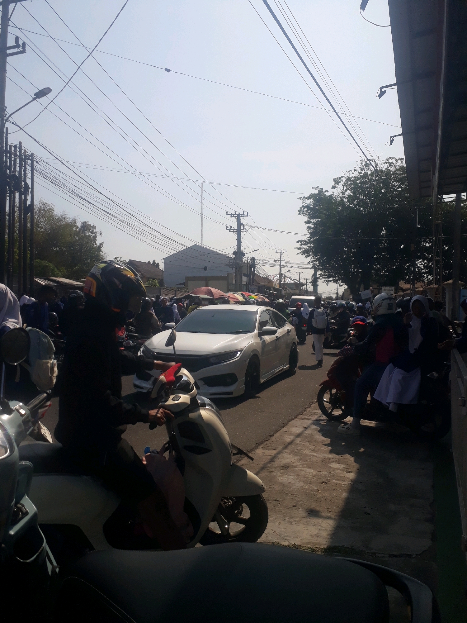 The image depicts a street scene in Pangkalpinang, Indonesia, characterized by a traffic jam and numerous pedestrians. A white sedan is prominently featured in the center, surrounded by a crowd of people on motorcycles. Many individuals are wearing helmets, indicating they are on motorcycles. The environment is urban, with buildings visible in the background and an overhead network of power lines. The sky is clear and bright, suggesting daytime. The foreground shows several motorcycles, some of which are partially obscured. The overall impression is one of a busy, congested street.