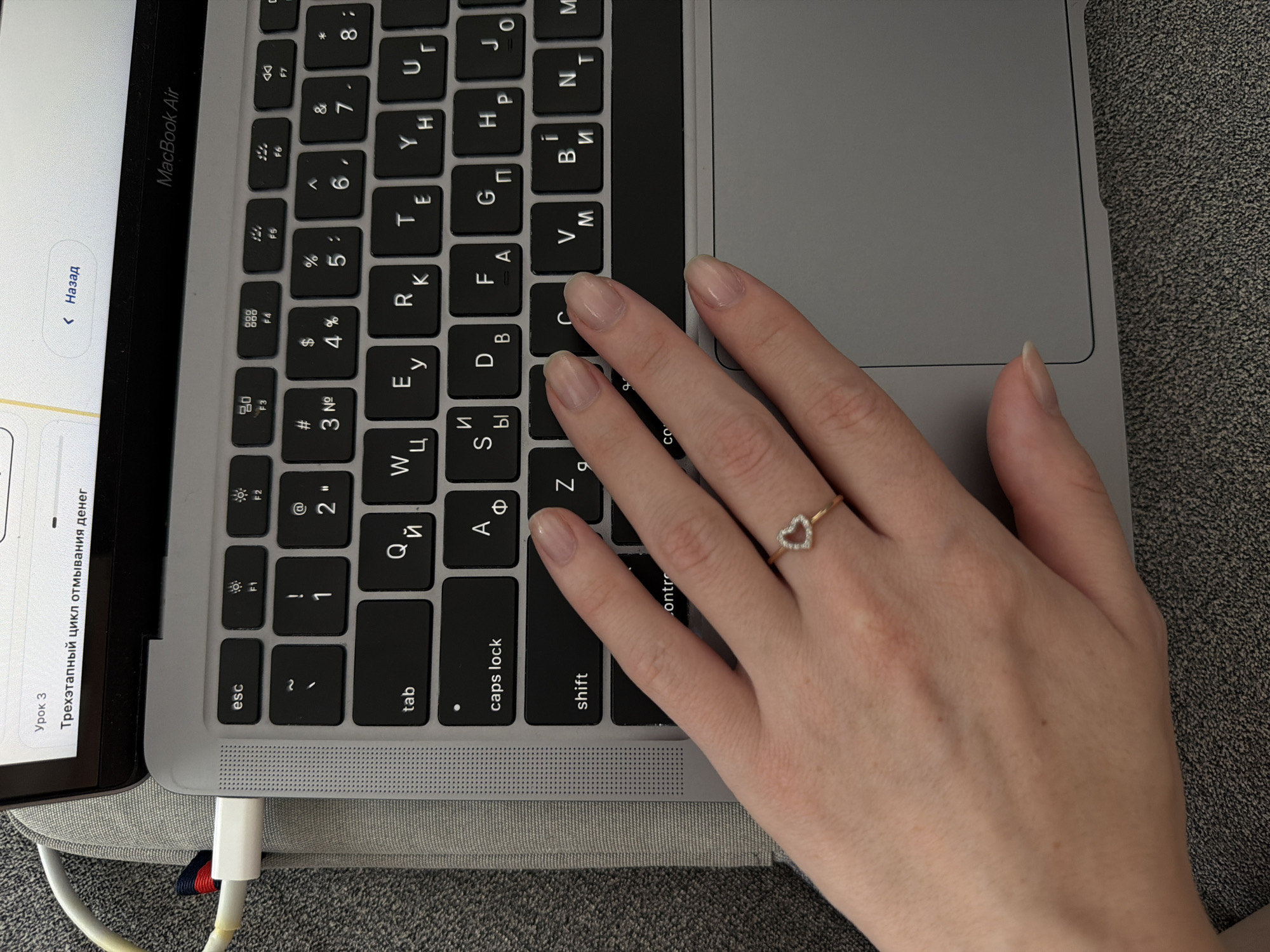 A close-up shot shows a person's left hand, adorned with a heart-shaped ring, resting on a laptop keyboard. The laptop appears to be a MacBook Air, identifiable by the "MacBook Air" logo on the screen bezel. The keyboard is illuminated by the screen's glow, with visible keys labeled in Cyrillic script, indicating it is likely a Russian or Ukrainian keyboard layout. The background consists of a textured gray surface, possibly a sofa or a laptop case, and a white USB-C cable plugged into the side of the laptop. The overall lighting suggests an indoor environment, possibly a home or office. There is no indication of the location being in Kyiv, Ukraine.