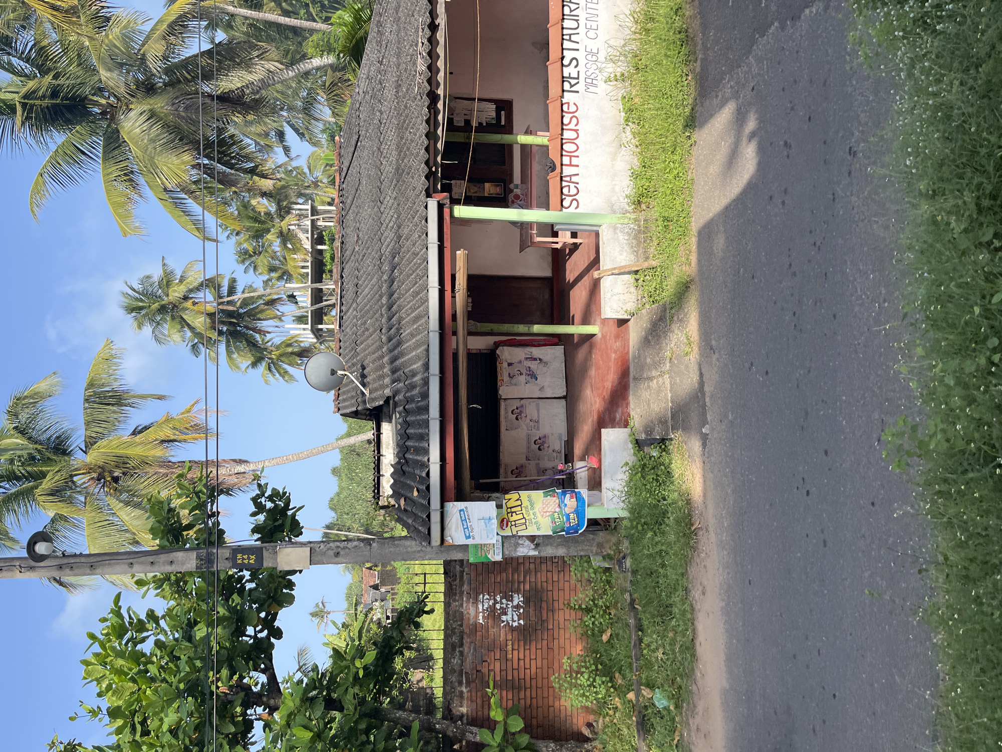 A low-angle, full outdoor shot captures the facade of a single-story building with a corrugated metal roof. The building is painted light pink and appears to be a commercial establishment, with "SEA HOUSE RESTAURANT MASSAGE CENTER" painted in red and black lettering on the lower part of the wall. A wide overhang shelters the entrance, which has two glass-paneled doors. Inside, there are two refrigerators or freezers. Various posters are displayed on the walls and on a utility pole to the left of the entrance. A brick wall borders the property on the left, with lush green vegetation growing along its base and in front of the building. Palm trees and other tropical foliage are visible in the background, suggesting a warm, possibly coastal, climate. The foreground features an asphalt road with sparse green grass and small white flowers along its edge. The sky above is clear and blue. The scene is static, with no people or animals visible. The overall impression is one of a small business in a tropical setting.