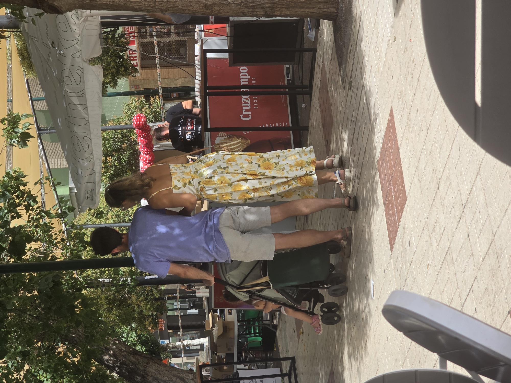The image captures an outdoor scene, possibly a street fair or market, in Málaga, Spain. In the foreground, a man in a purple t-shirt and grey shorts pushes a stroller with a young child sitting in it. To his right, a woman in a yellow floral dress walks beside him. Behind them, a man wearing a black t-shirt with text and graphics is visible. Various stalls and structures are present, including a white umbrella with "Heineken" branding and a red banner with "Cruzcampo" text. The ground is paved with light-colored tiles. Trees and foliage are visible in the background, suggesting a sunny day.