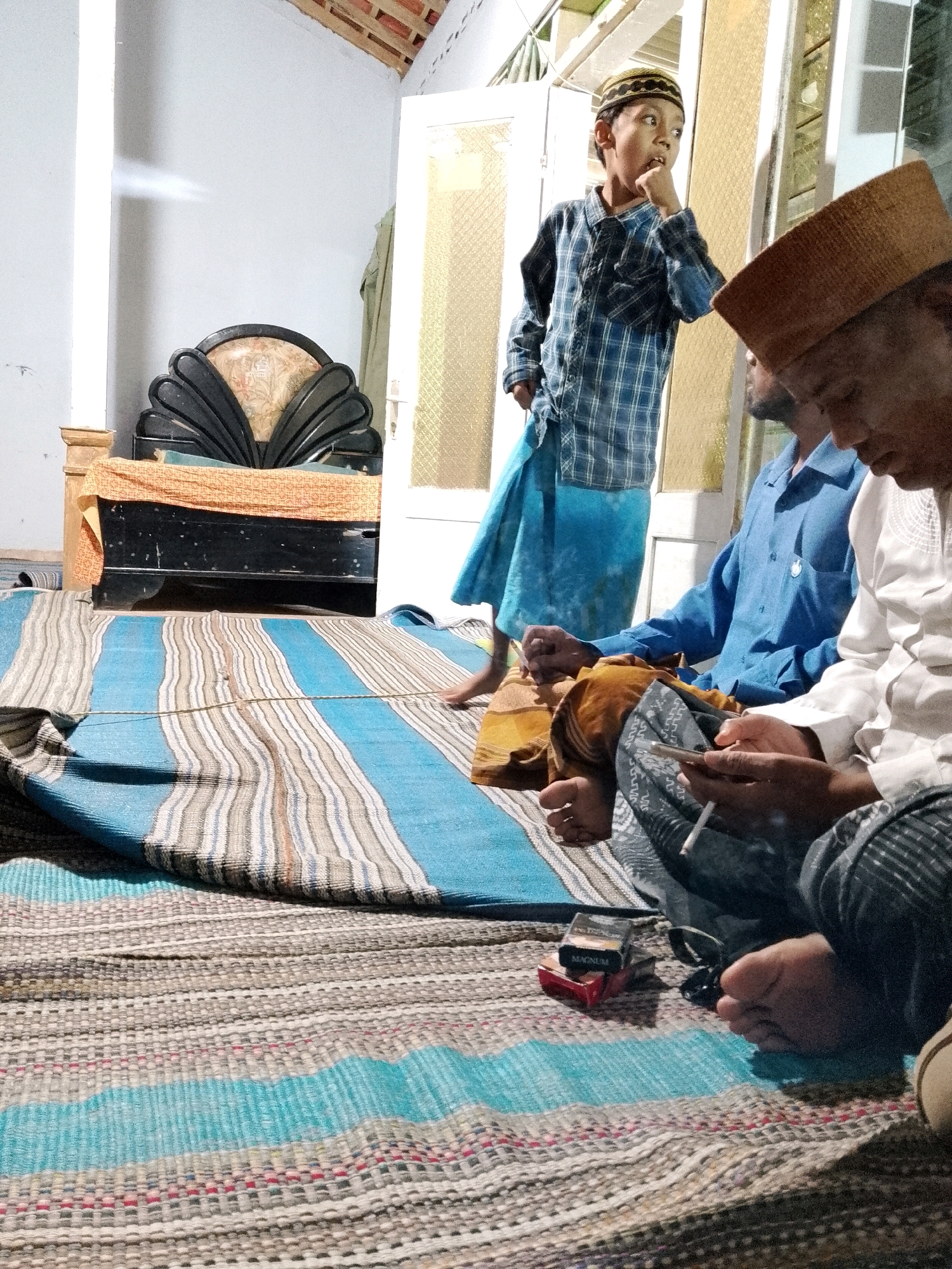 In a room in Rejoyoso, Indonesia, a group of individuals, likely male, are gathered on the floor, seated on woven mats. One young person, wearing a blue plaid shirt and a patterned peci (a type of cap), stands and appears to be looking off to the side, with a hand near their mouth. Two older individuals are seated closer to the foreground, also wearing pecis. One is wearing a blue shirt and the other a white shirt. Both are holding cigarettes, and one is looking at a mobile phone. A pack of "MAGNUM" brand cigarettes is visible on the mat near them. The environment appears to be a simple indoor setting with a white-painted wall and exposed wooden ceiling beams. A large, ornate headboard of a bed or sofa is visible in the background, draped with orange fabric. The floor is covered with a large, striped woven mat with blue and grey stripes.
