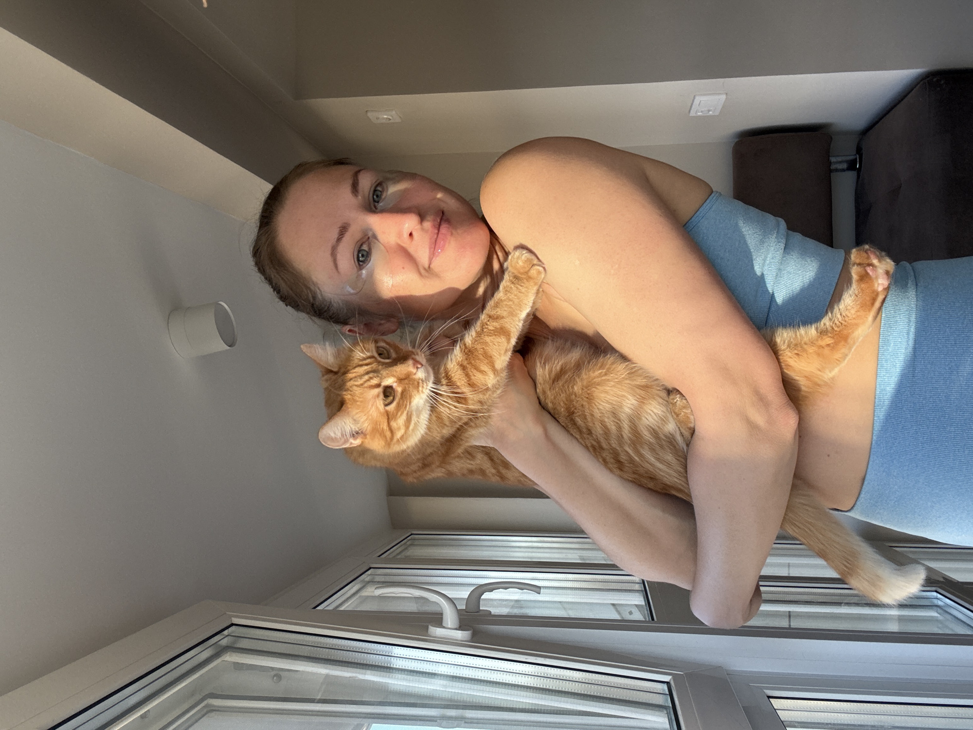 A woman is holding an orange tabby cat in front of a window. The woman is wearing a light blue top and has a bandage on her face. The location is Chaikovsky, Russia.