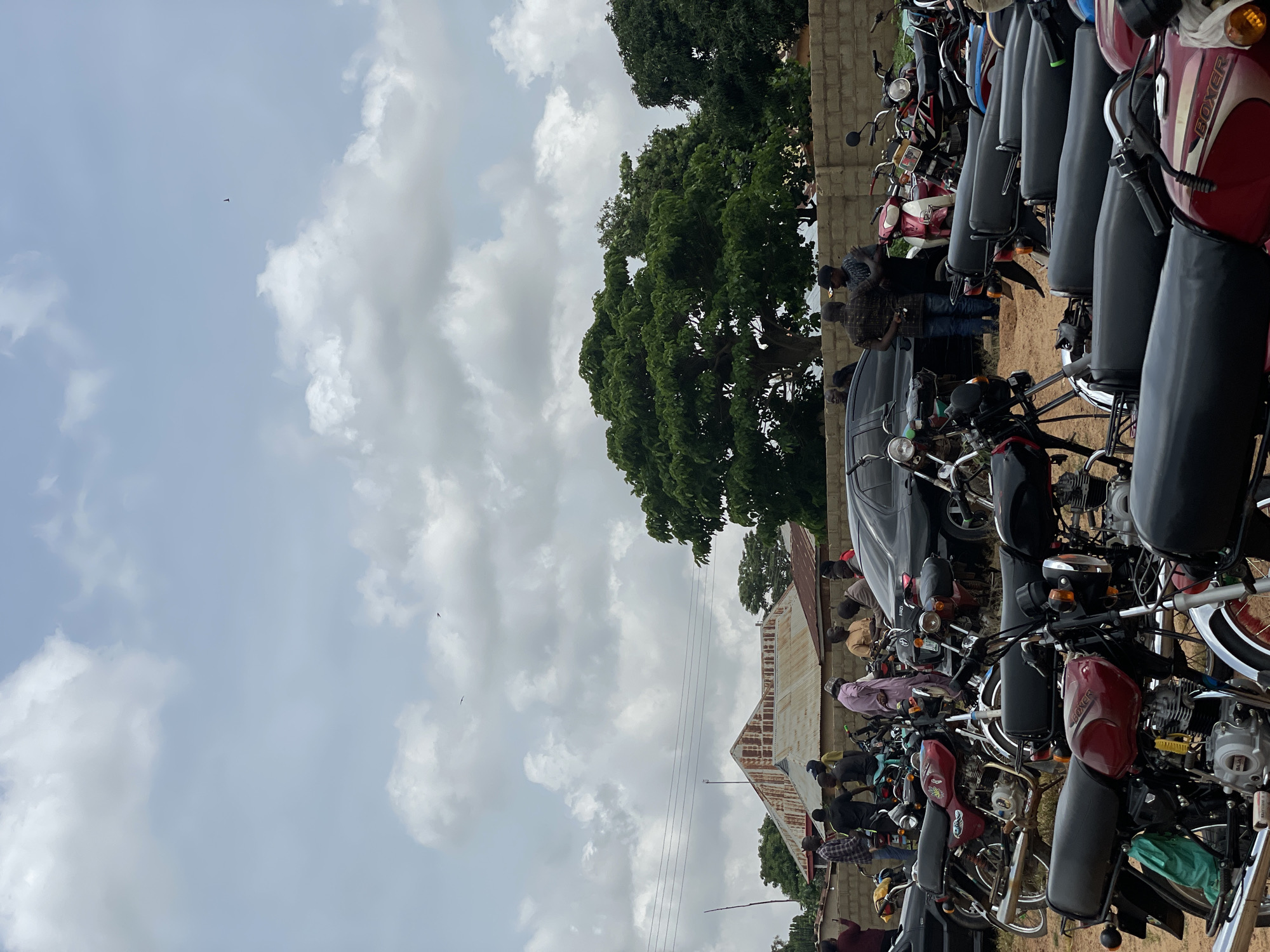 In Kassa, Nigeria, a gathering of people and parked motorcycles unfolds under a cloudy sky. In the background, a building with a rusty metal roof stands against the horizon, while lush green trees punctuate the landscape. The foreground is dominated by a collection of motorcycles, some red and black, parked closely together on a sandy surface. Several individuals are present, some standing near the motorcycles, others engaged in conversation. A black sedan is also visible, parked amongst the motorcycles. The scene suggests an informal outdoor meeting or event, possibly a community gathering or a market day.
