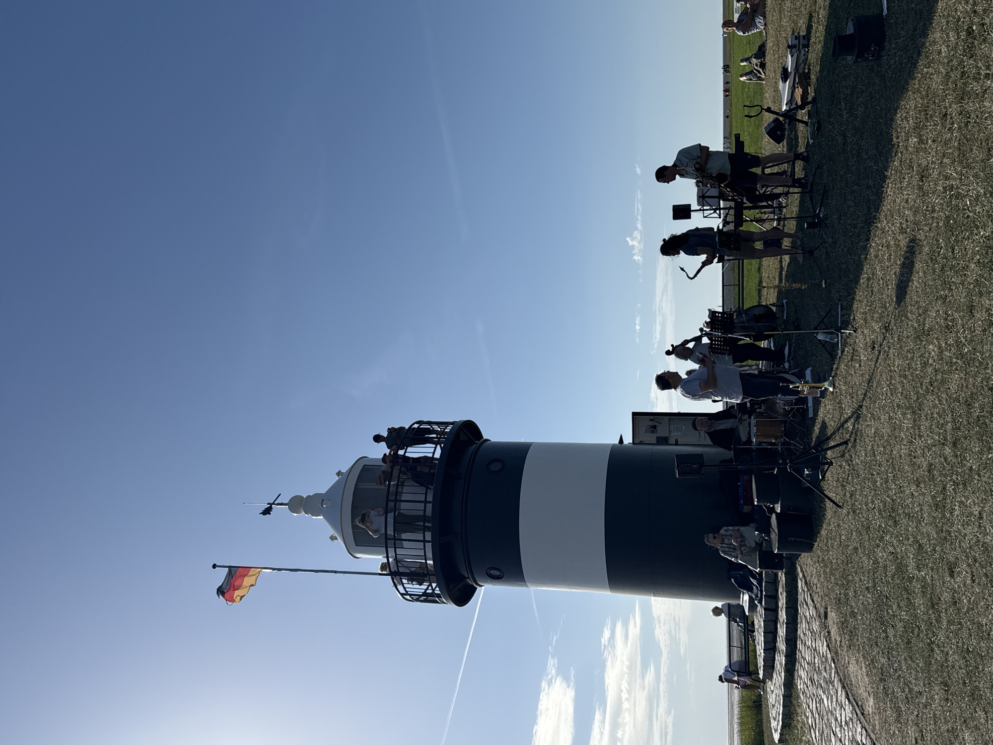 This is a vertical, medium-shot, eye-level, outdoor photograph taken on a sunny day. The main subject is a black and white striped lighthouse, which is on the left side of the frame. A German flag flies from a flagpole to the left of the lighthouse. There is a group of people on a viewing platform at the top of the lighthouse. In the foreground, there is a band playing music. One person plays a saxophone, another plays a bass guitar, and a third plays drums. There are also some spectators sitting on the grass, watching the band. The environment appears to be a grassy area near the coast, with a clear blue sky overhead.