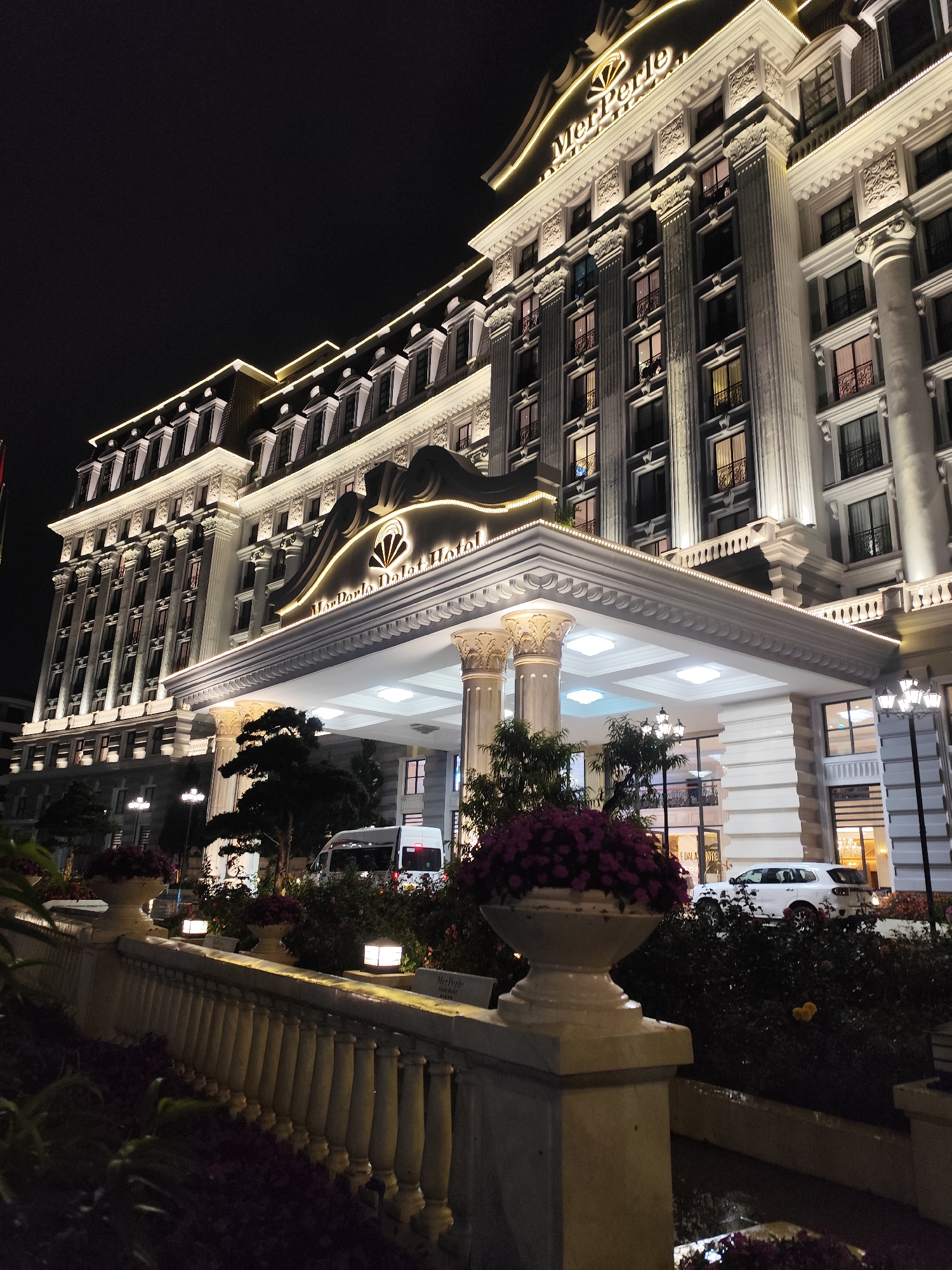 The image displays the grand, illuminated exterior of the MerPerle Dalat Hotel at night, located in Da Lat, Vietnam. The multi-story building showcases a classical architectural style, featuring numerous prominent columns, ornate cornices, decorative balconies, and a distinctive mansard roof outlined with glowing golden lights. The entire facade is brightly lit with white and warm yellow lights, highlighting its intricate details.
The hotel's name, "MerPerle Dalat Hotel," is clearly visible in illuminated script on the entrance canopy, accompanied by a stylized shell or fan logo. The word "MerPerle" with the same logo is also present near the top of the building.
In the foreground, a stone balustrade with ornate posts curves upwards, bordering landscaped garden beds. These beds are filled with dark foliage and potted plants bursting with vibrant purple flowers. Various outdoor lamps and streetlights provide additional illumination to the pathways and surrounding area. Under the expansive, well-lit entrance canopy supported by large columns, a white van and a white SUV are partially visible, indicating vehicle activity at the hotel's entrance. The sky is dark, accentuating the hotel's impressive and luxurious appearance against the clear night. There are no people clearly visible or specific activities taking place, but the scene suggests the hotel is open and operational.