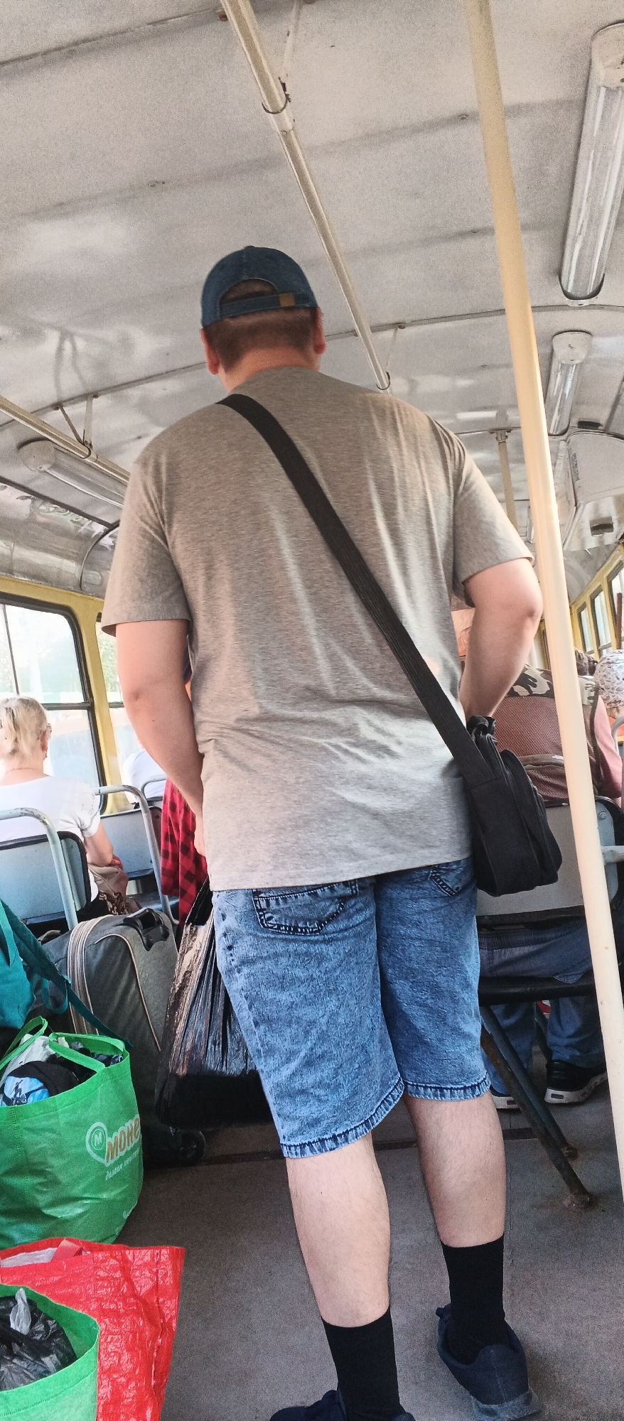 The image depicts the interior of a public transportation vehicle, likely a bus or tram, in Izhevsk, Russia. The perspective is from behind a man wearing a grey t-shirt, blue denim shorts, black socks, and blue sneakers. He has a blue baseball cap on and a black messenger bag slung across his body. Beside him are several bags, including a green reusable shopping bag with the logo "MORE" and a red plastic bag. In the background, other passengers are visible, including a woman with blonde hair facing away from the camera and another passenger wearing a patterned headscarf. The vehicle's interior features metal poles, fluorescent lighting fixtures on the ceiling, and yellow-tinted windows. The flooring appears to be a light-colored composite material. The overall scene suggests a casual daytime journey.