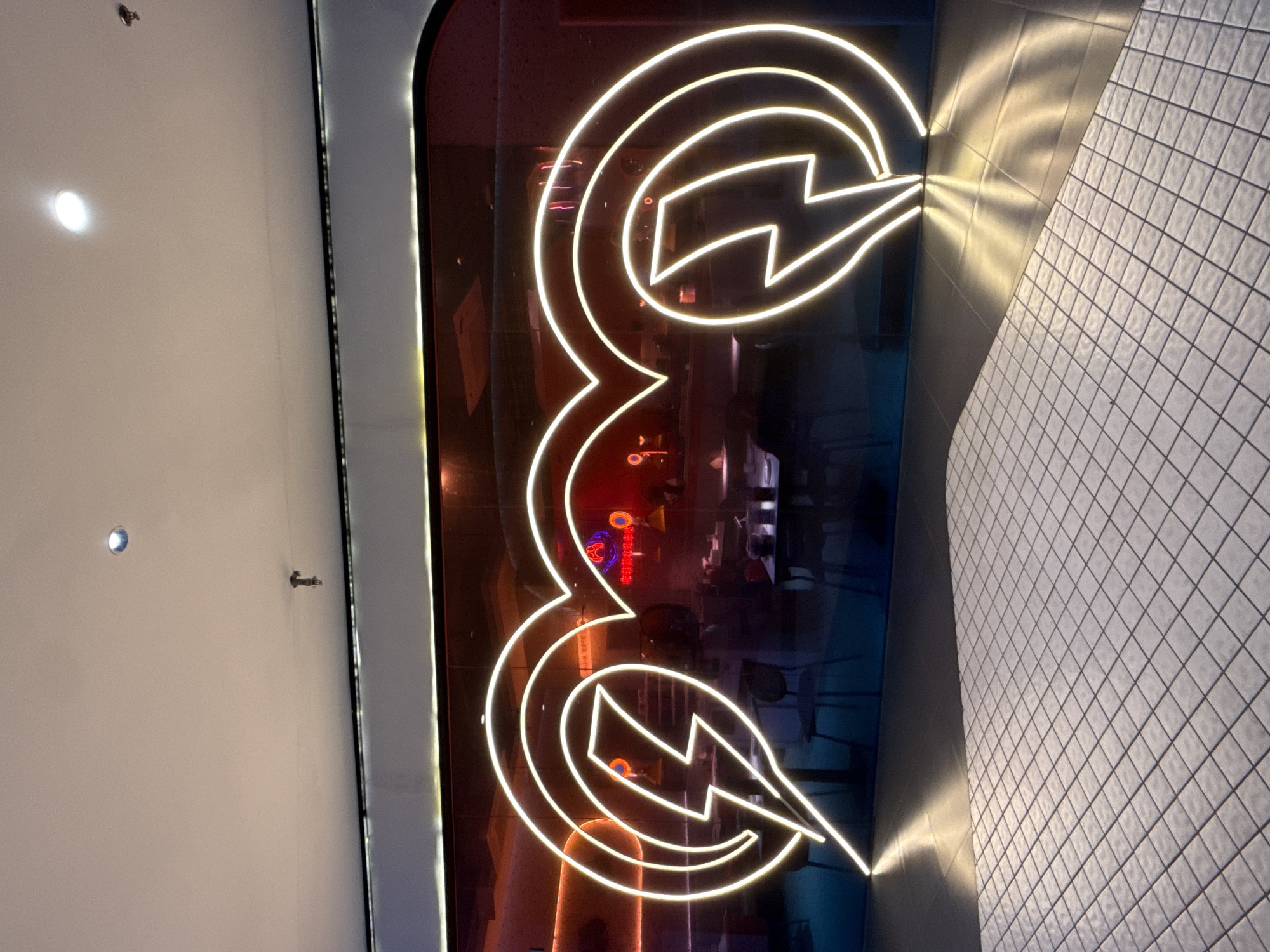 The image depicts a neon sign shaped like two intertwined lightning bolts forming abstract heart-like shapes. The neon sign is bright white and appears to be mounted on a dark, reflective surface that is curved at the top. Behind the neon sign, a restaurant interior is visible through the glass. This restaurant has red and blue lighting, with tables and chairs set up, suggesting it is open for business. The floor in the foreground is tiled with small white squares arranged in a diamond pattern, reflecting the light from the neon sign. The scene appears to be indoors, possibly in a shopping mall or a commercial district. The location is Huizhou, China.