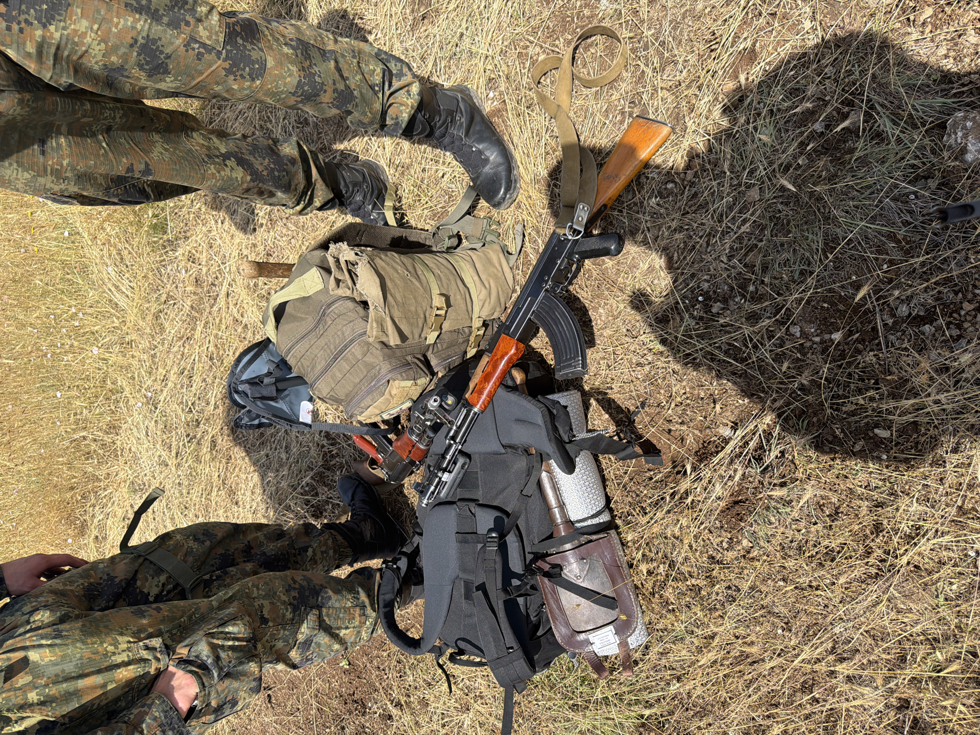Two individuals, wearing camouflage uniforms, stand on dry, grassy ground. Their lower legs and feet are visible, along with their tactical boots. Scattered on the ground are two backpacks, one a green canvas rucksack and the other a black tactical backpack. A rifle, appearing to be an AK-variant, lies on the ground alongside the backpacks. One of the backpacks has a cylindrical object with a leather casing attached to it. The environment is outdoors, with dry vegetation and dirt covering the ground. The location is identified as Beliakovets, Bulgaria.